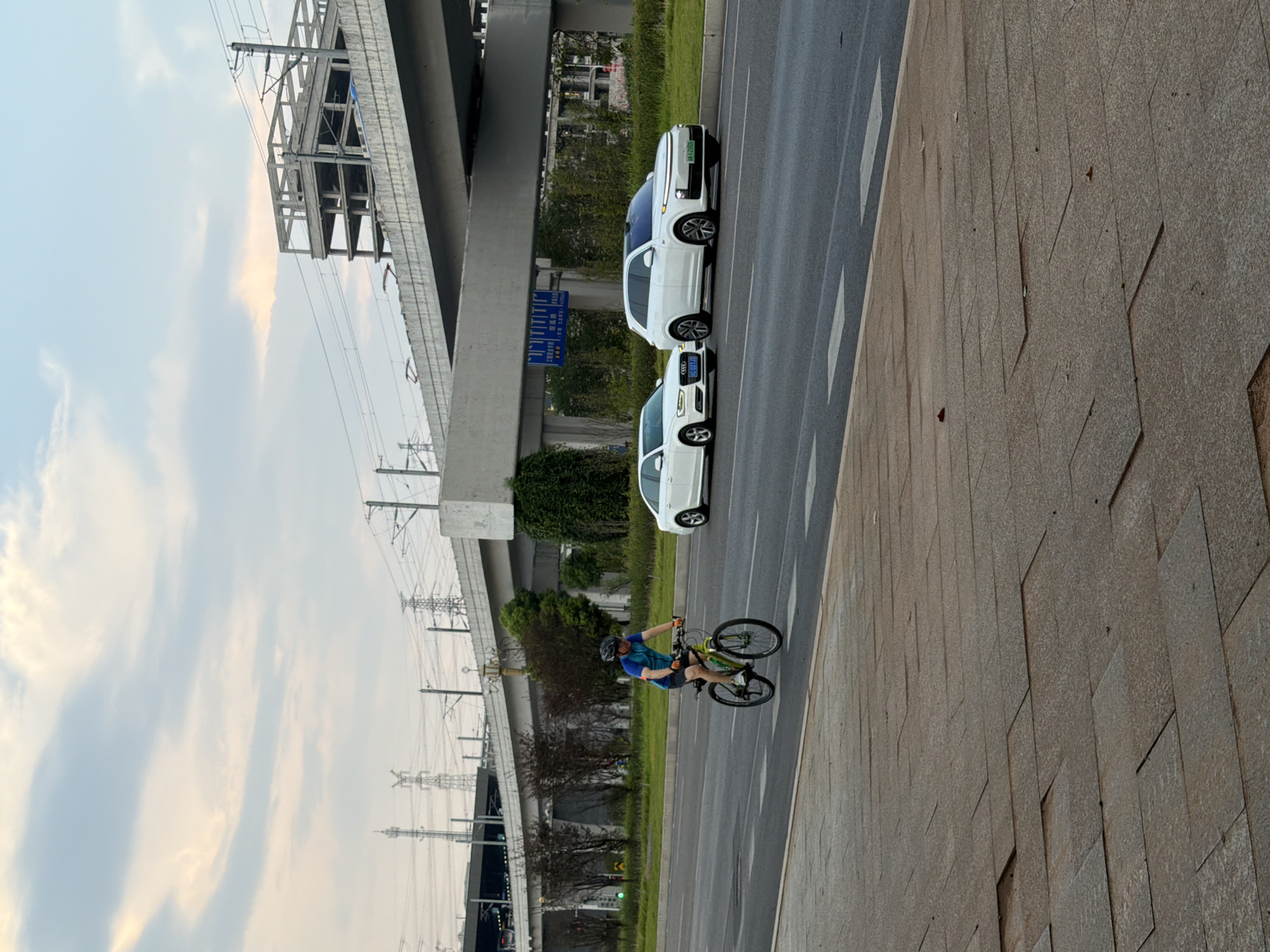 In Jiangjin, China, a person wearing a blue and yellow t-shirt, black shorts, and a helmet cycles on a paved path next to a road. The cyclist is on a green bicycle and appears to be moving from left to right. White cars are driving on the road in the same direction. In the background, a large elevated highway structure looms over the scene, with industrial buildings and power lines visible further beyond. The sky is partly cloudy with hints of sunlight. The foreground shows a tiled sidewalk.