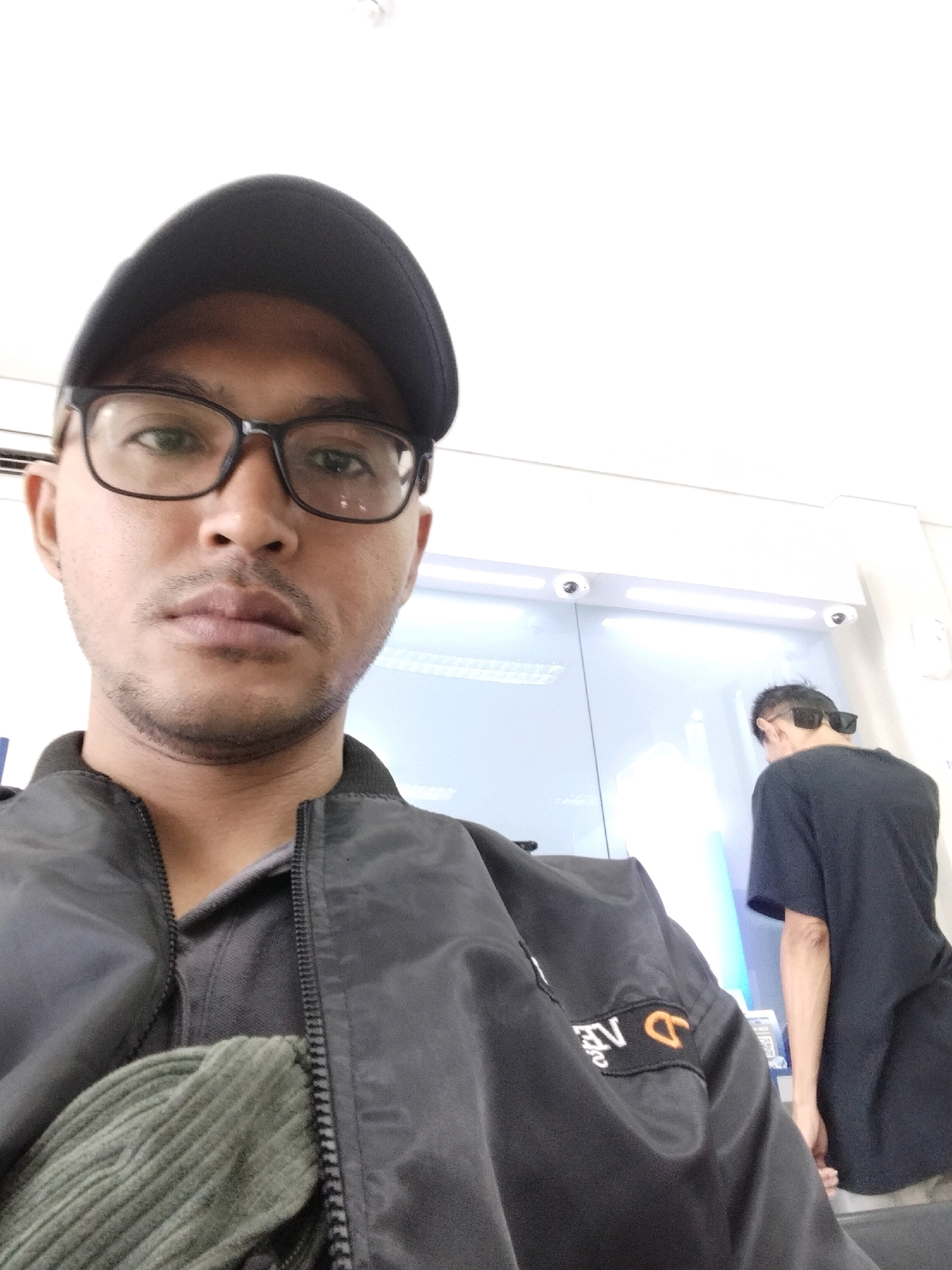 In this selfie taken from a close-up perspective, a man wearing a black baseball cap and glasses is in the foreground. He has a mustache and a slight goatee, and is wearing a black jacket over a grey polo shirt. A green ribbed bag is partially visible under his jacket. In the background, another person wearing a black t-shirt and sunglasses is facing away from the camera, interacting with an ATM machine. The environment appears to be an indoor commercial space, possibly a bank or a convenience store, with bright fluorescent lighting and security cameras visible. This scene is situated in Kuta, Indonesia.