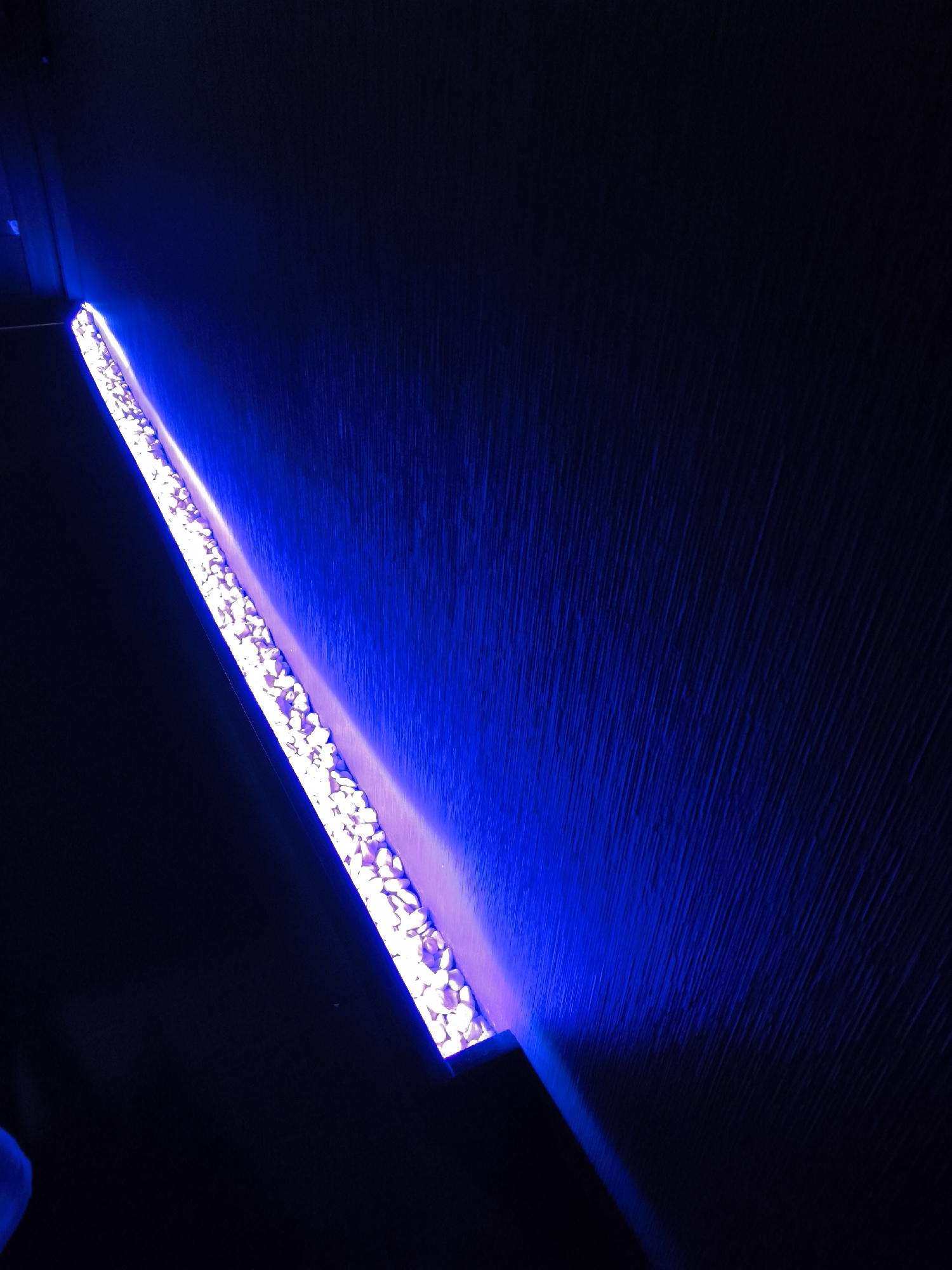 A dimly lit, angled view shows a water feature, possibly a waterfall, illuminated with intense blue light. The water cascades from a rectangular trough filled with small, white stones. The background appears to be a textured wall, also illuminated by the blue light, which accentuates its surface. The scene's location in Moscow, Russia, is not discernible from the image. No people or interactions are visible.