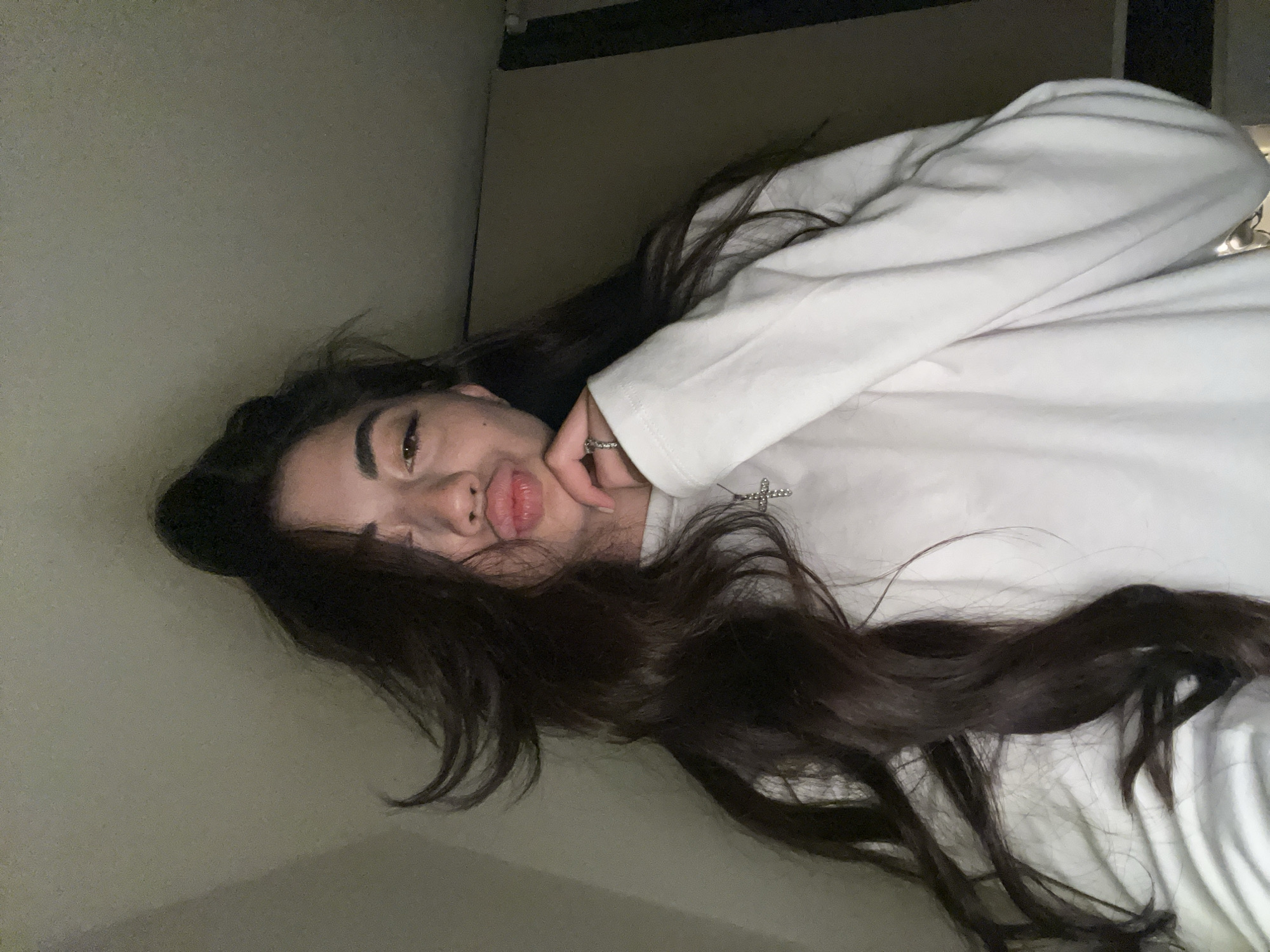 A young woman with long, dark, wavy hair is captured in a selfie. She is wearing a white long-sleeved shirt with a silver cross necklace. Her right eye is open and looking at the camera, while her left eye is partially closed. Her lips are pursed in a kiss-like gesture, and her right hand is raised to her chin. The background appears to be an indoor setting with plain walls. The lighting is dim, casting shadows on the walls. The overall mood is casual and playful. The image does not contain any identifiable features or context that would allow for determination of its location in Chotěšov, Czechia.