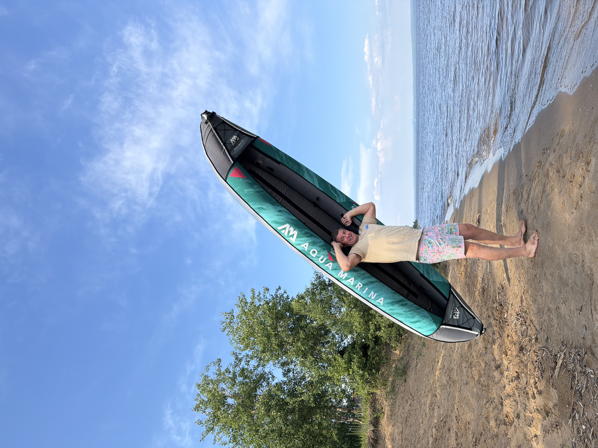 An adult male stands on a sandy shore, holding an inflatable paddleboard over his head. The paddleboard is green and grey and has "Aqua Marina" written on it. The man is wearing a tan t-shirt and patterned shorts. He is barefoot. The background shows a large body of water with a few trees and a blue sky with some clouds. This scene is likely the Kama River embankment in Chaikovsky, Russia, given the presence of a large river and the specific model of the paddleboard, which is popular in Russia.