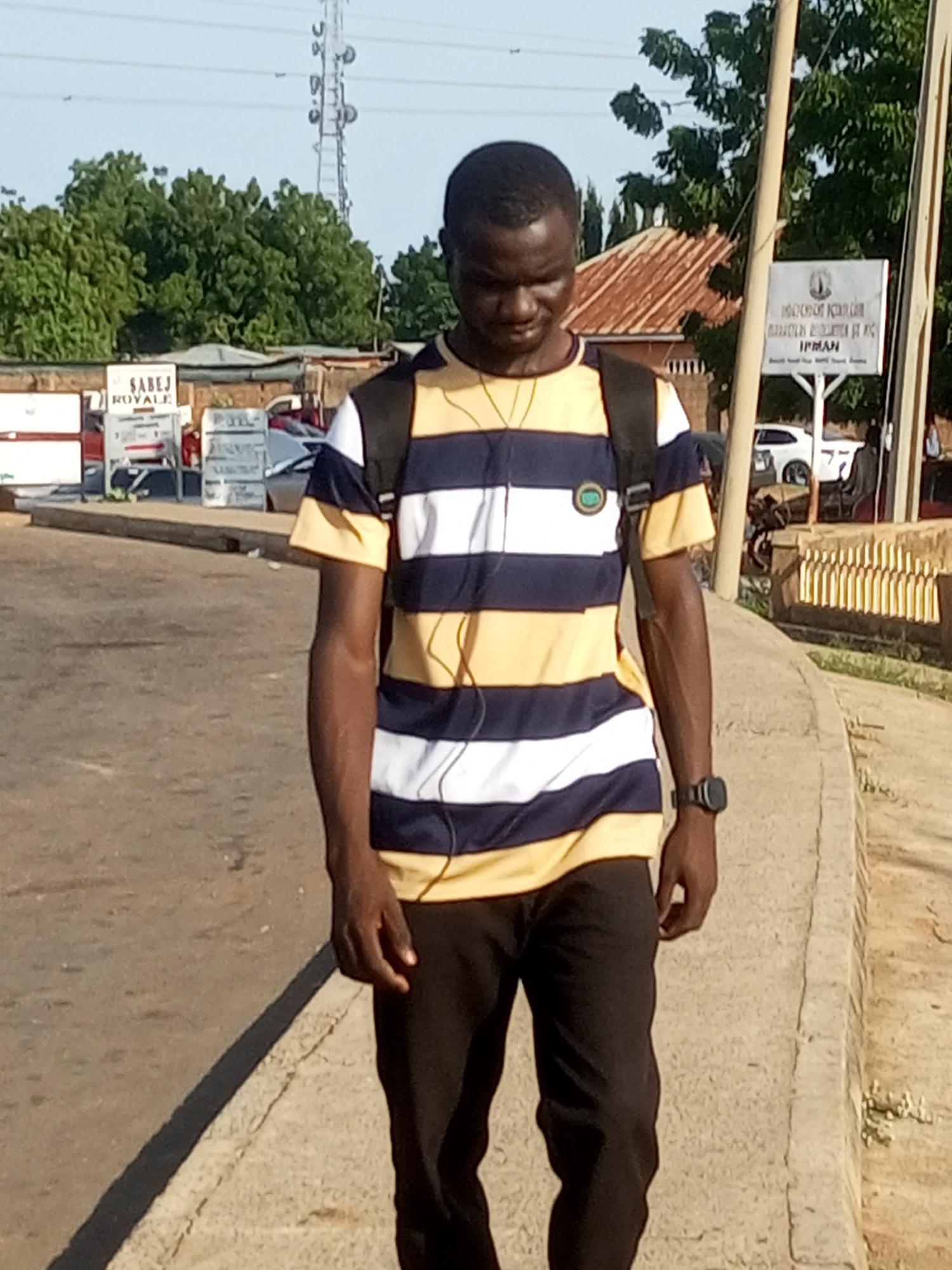 In Gombe, Nigeria, a young Black man with dark skin and short hair walks down a paved pathway next to a road. He is wearing a horizontally striped t-shirt with alternating bands of navy blue, white, and yellow, and dark pants. A black backpack is slung over his shoulders. He wears a wristwatch on his left wrist and earbuds in his ears. To his left, a sign reading "SABEJ ROYALE" is visible. In the background, there are trees, a rusty-roofed building, a tall telecommunications tower, and a white sign with text and a logo. A white car is parked on the side of the road. The pathway is bordered by a concrete curb.