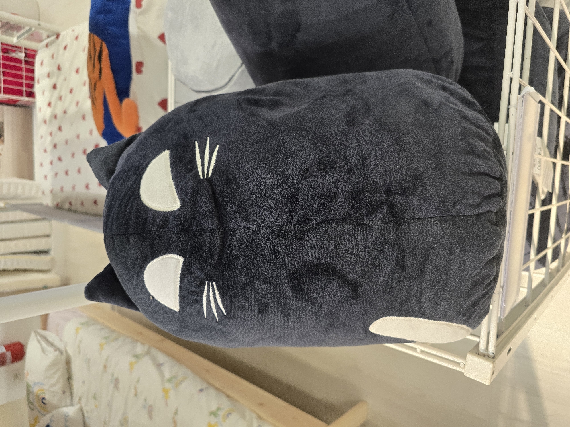 A dark blue plush cat toy is prominently displayed on a white wire shelf in what appears to be a retail environment. The toy is large and ovular, with white embroidered eyes and whiskers. To the left of the cat toy, a child's bed with patterned bedding and a pillow can be seen. In the background, more beds and shelves are visible, suggesting a children's bedding or toy store. The lighting is bright and even, typical of an indoor retail space. There are no people visible in the image. The location is identified as Baronissi, Italy, but this cannot be visually confirmed from the image alone.