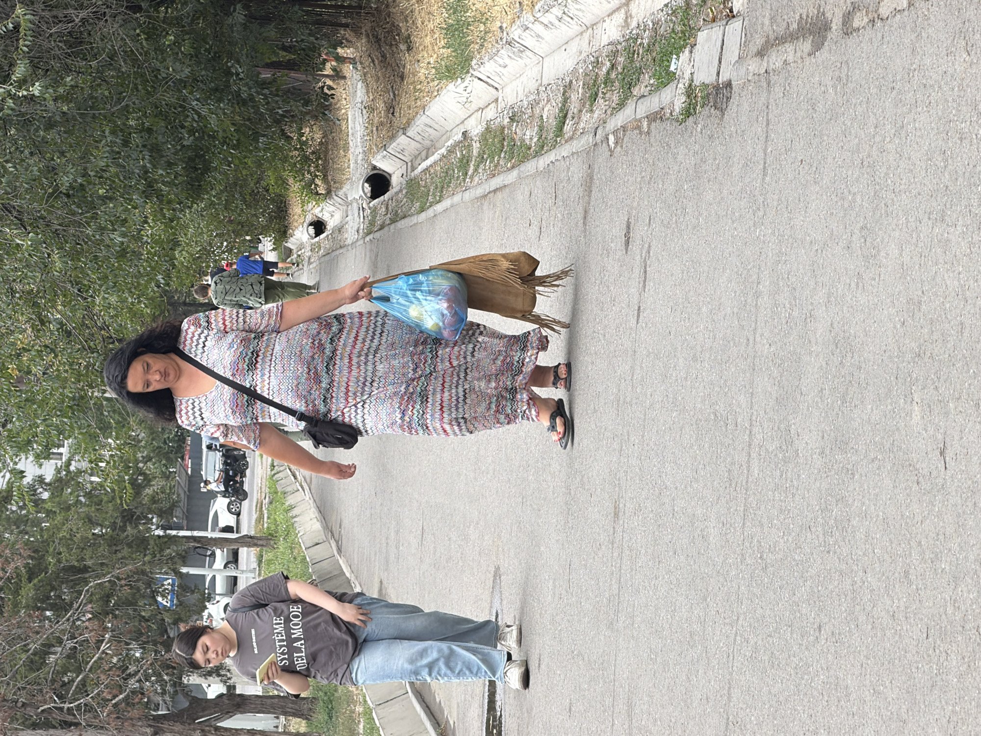 An outdoor medium shot captures two women walking along an asphalt pathway in a city. The woman in the foreground, on the right, is wearing a long, multicolored patterned dress and dark flip-flops. She carries a blue plastic bag filled with items and a brown suede fringed bag over her left shoulder. The woman in the background, on the left, is wearing a gray t-shirt with text and light blue jeans. She holds a smartphone in her left hand and has a dark bag slung over her shoulder. Both women are walking away from the camera. The pathway is bordered by a concrete drainage ditch on the right and sparse vegetation and trees on the left. In the background, several vehicles and other pedestrians are visible. The weather appears to be overcast.