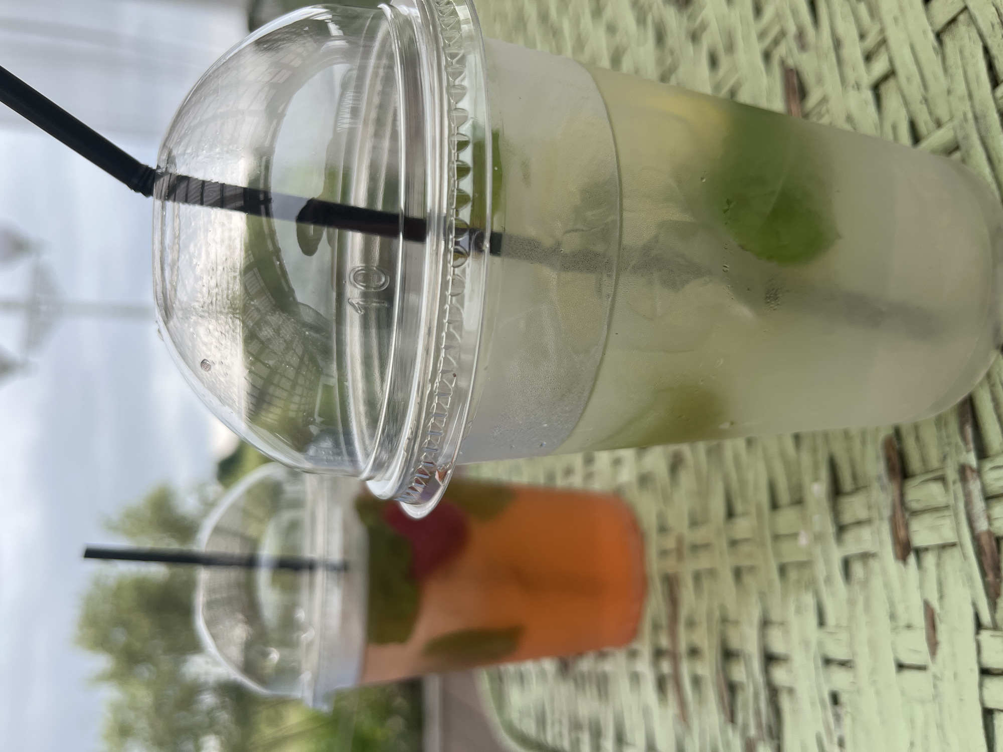 Two clear plastic cups with domed lids and black straws are placed on a light green woven surface. The cup in the foreground contains a light yellow beverage with ice and green mint leaves. The cup in the background contains a pink or orange beverage with ice, mint leaves, and possibly a red fruit. The background is blurred, showing green foliage and a cloudy sky, suggesting an outdoor setting. The scene appears to be in the city of Severynivka, Ukraine, possibly at an outdoor cafe or seating area.