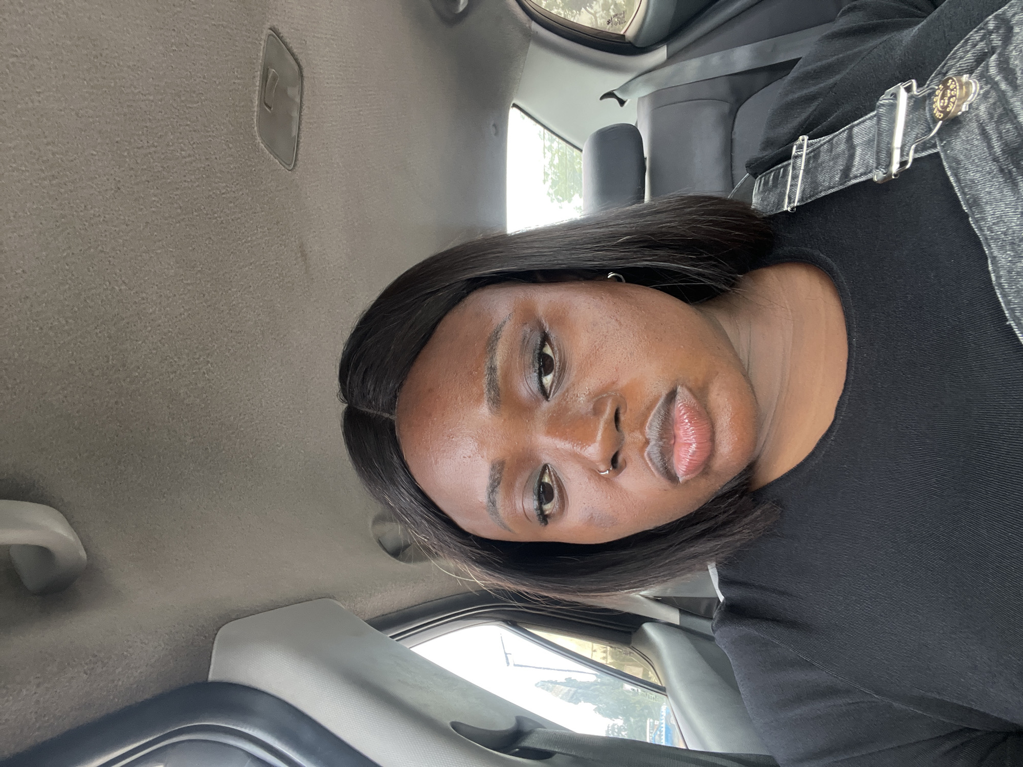 A young Black woman with dark, straight hair and full lips is photographed from the chest up, seated inside a vehicle. She is wearing a black t-shirt and denim overalls. Her gaze is directed downwards, and her facial expression appears somber. The interior of the car is visible, with the ceiling, headrest, and seatbelt. The lighting suggests it is daytime, and through the window, a glimpse of trees and buildings can be seen, indicating an outdoor setting. This photograph was taken in Galadimawa Village, Nigeria.