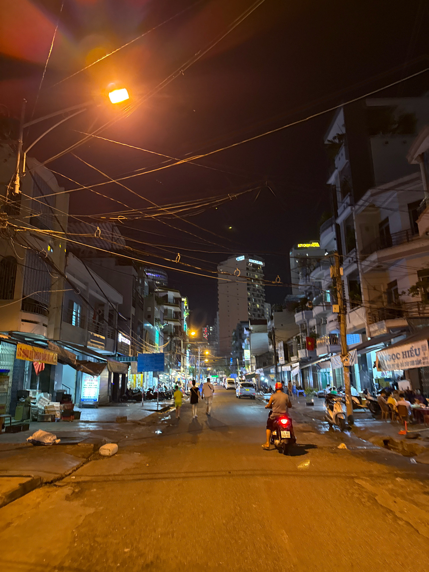 The image depicts a nighttime street scene in Nha Trang, Vietnam, characterized by a busy urban environment with numerous buildings, overhead wires, and a lit streetlamp. The street is paved and appears wet, possibly from rain. Several individuals are present: a person riding a motorcycle is prominently featured in the foreground, facing away from the camera. Further down the street, a group of people are walking, and others are seated at outdoor tables, likely at a restaurant or cafe. The buildings lining the street are a mix of multi-story structures, some appearing to be hotels or apartment buildings, with various signs and advertisements displayed. The overall atmosphere is one of a lively, commercial street at night, illuminated by artificial lighting.