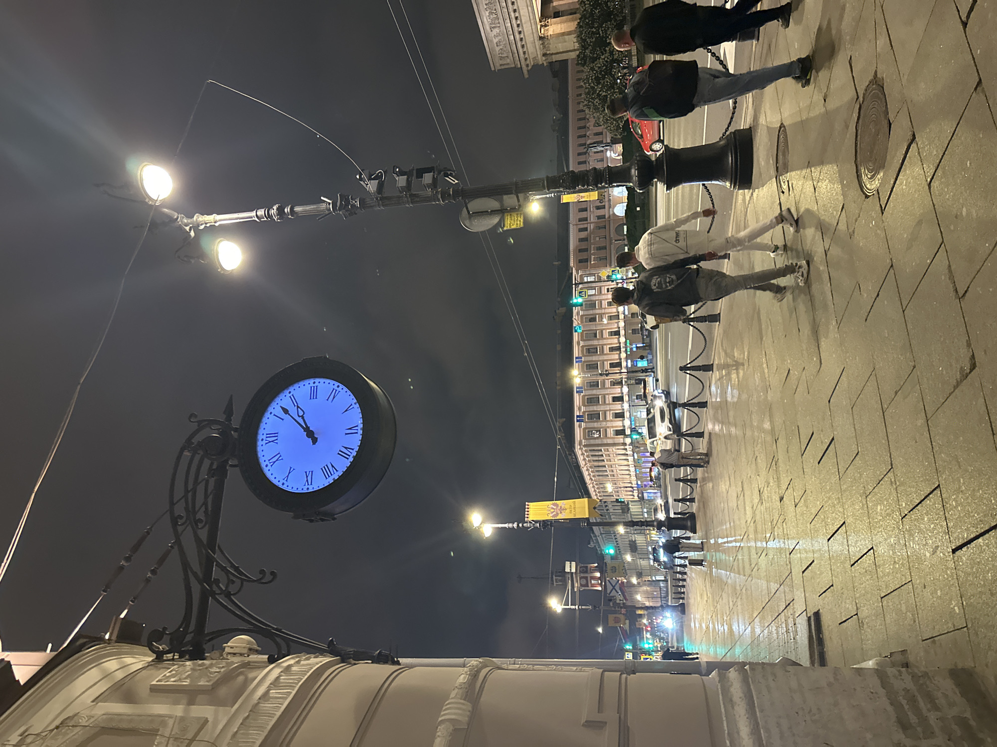 The image captures a night scene in Saint Petersburg, Russia, featuring a prominent street clock affixed to a building with classical architectural elements. The clock face is illuminated with Roman numerals. Above it are two street lamps. The perspective is from ground level, angled upwards towards the clock and street. Along the sidewalk, multiple pedestrians are walking, some appearing to be in groups. The street is lit by numerous streetlights, extending into the distance, where buildings line both sides of the street. A yellow banner hangs between the streetlights. The ground appears wet and reflective.