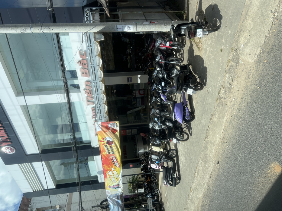 A multistory Yamaha dealership displays its logo prominently. Awnings shade the entrance. Various motorcycles are parked in a row along the sidewalk. Banners advertise promotions, while utility poles with wires rise along the building. The image seems to have been taken from an elevated perspective.