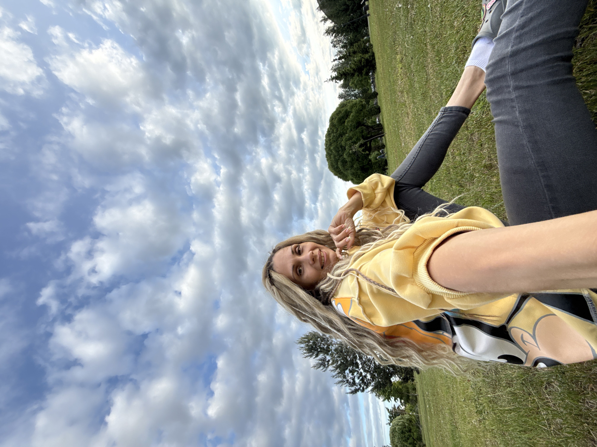 A young woman with long, light blonde hair, appearing to have dreadlocks or braids, is captured in a selfie taken outdoors. She is smiling directly at the camera, conveying a cheerful and relaxed mood. She is wearing a bright yellow long-sleeved top or hoodie, which features a partial graphic of a cartoon character's face, possibly Mickey Mouse, with a prominent rounded ear visible. Her lower body is clad in dark gray or black jeans, and white socks are visible above her shoes.
The setting is a vast green expanse of grass, suggesting a park or an open field. In the background, numerous trees with dark green foliage are visible, including one particularly large, rounded tree on the right side of the frame. The sky above is partly cloudy, with a mix of blue patches and fluffy white and gray clouds. The overall lighting suggests it is daytime, possibly late morning or afternoon. The image is oriented sideways, implying the person was reclining or lying down when taking the selfie, looking up at the sky and surrounding environment. The location is contextualized as Заволжье, Russia, depicting a pleasant outdoor scene in that region.