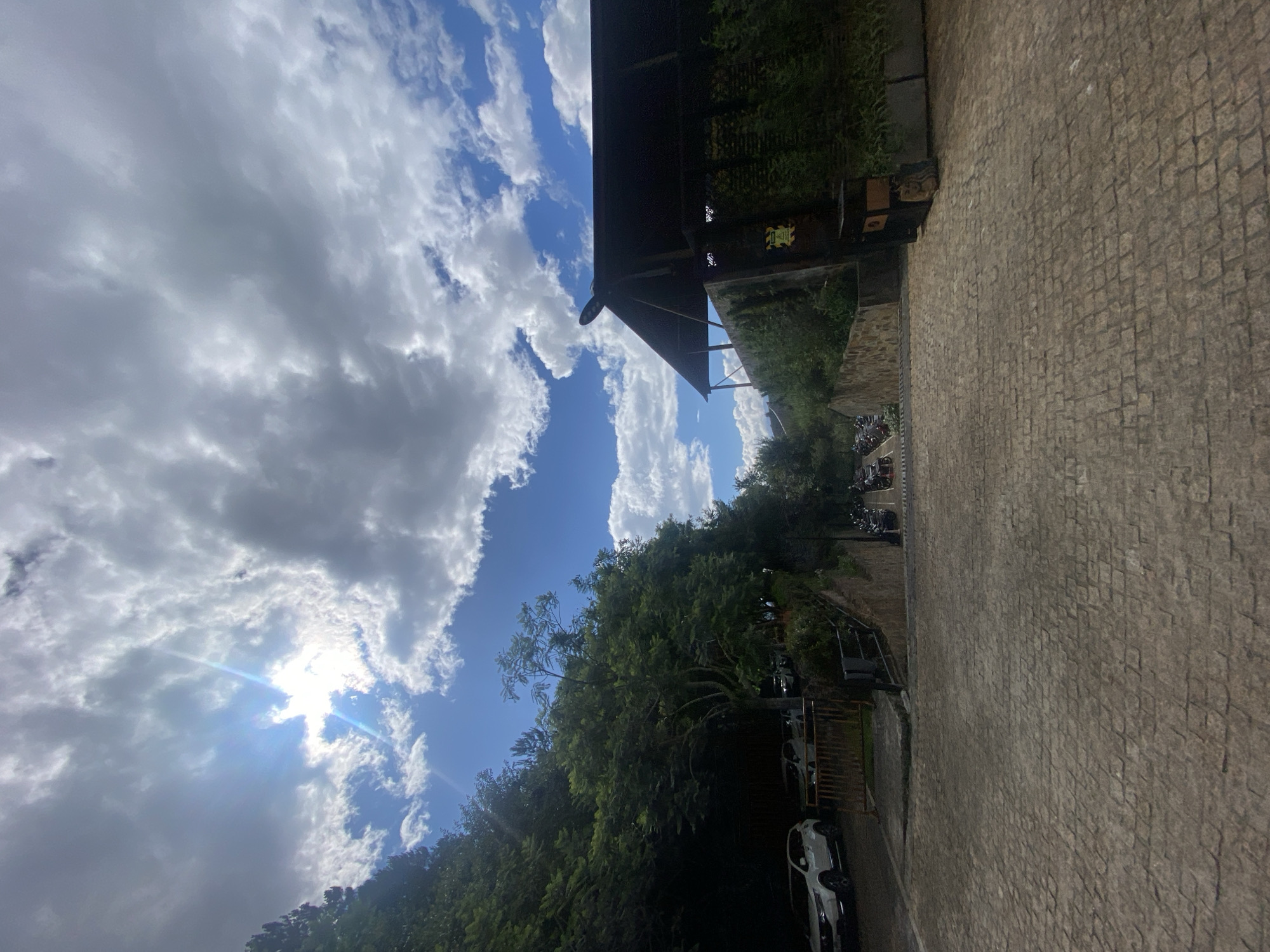 This wide-angle outdoor photograph depicts a descending cobblestone pathway flanked by dense greenery, parked vehicles, and architectural structures, located in Púng Luông, Vietnam.
The upper portion of the image showcases a bright blue sky with numerous white cumulus clouds of varying density. A prominent sun flare with distinct rays originates from the upper-left quadrant, where the sun is partially obscured by clouds, indicating direct sunlight.
On the left side, a steep, densely vegetated hillside is covered with a thick canopy of dark green trees and foliage. Below this natural incline, on a paved surface, two white sport utility vehicles are parked. A low, reddish-orange metal barrier separates this parking area from the main cobbled path.
The right side features a building with a distinctive dark, multi-angled roof, exhibiting characteristics of traditional regional architecture. The lower facade of this structure and an adjacent retaining wall are heavily overgrown with lush green climbing plants or shrubs. A yellow and black warning sign is affixed to this wall, and a dark rectangular object, possibly a planter, with a framed image leaning against it, is situated near its base.
The central and foreground area consists of a wide, descending pathway paved with irregularly shaped, light grey-brown cobblestones, whose texture is clearly visible. Further down the slope, at the bottom-center and right, a line of dark-colored motorcycles or scooters is parked along the side of the path, partially obscured by shadow. No human figures are present in the scene.