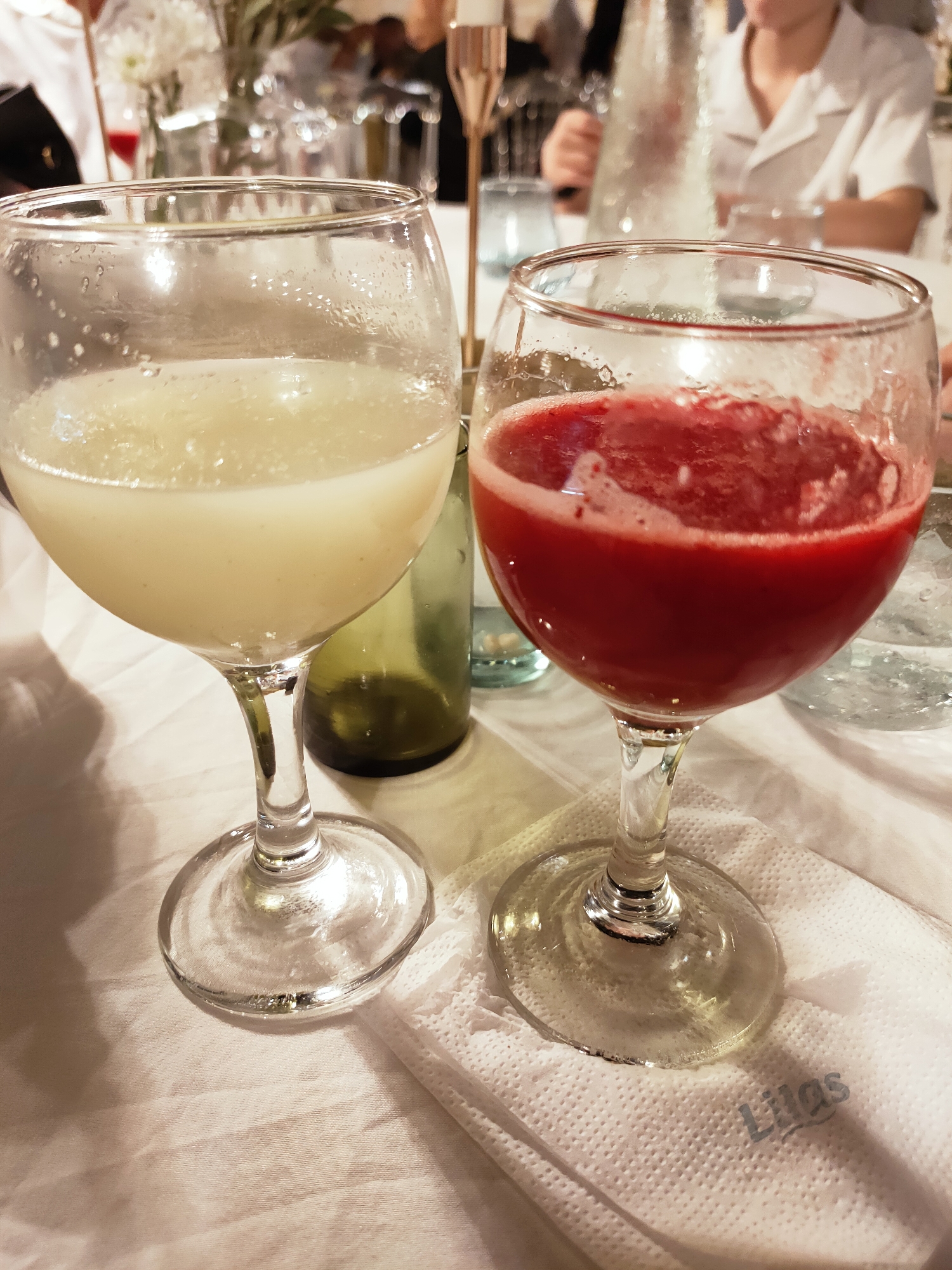 Two wine glasses containing beverages are placed on a white tablecloth. The glass on the left contains a light-colored liquid, possibly a smoothie or juice, while the glass on the right contains a vibrant red liquid, likely a fruit juice or cocktail. Both glasses are stemmed and have a wide bowl. A light-colored, textured napkin with the word "Lilas" embossed in blue is placed under the base of the right glass. In the background, blurred figures and table settings suggest a social gathering or a restaurant setting. The environment appears to be indoors, with soft lighting. No specific location in Cebalat, Tunisia is identifiable from the image itself.