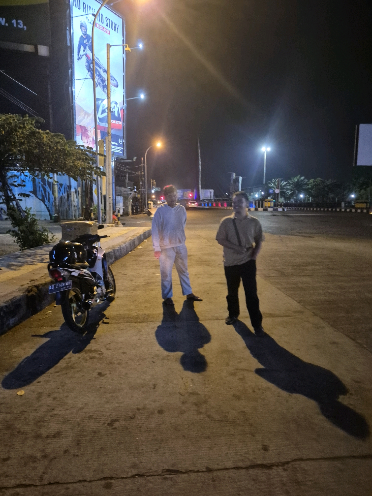 A nighttime street scene in Surakarta, Indonesia, features two men standing on a concrete surface near a parked motorcycle. A large billboard with a picture of a motorcyclist and the words "NO RIDE NO STORY BOLD" is visible in the background, illuminated by streetlights. The man on the left wears a light-colored hoodie and pants, while the man on the right is dressed in a beige collared shirt and dark pants. The motorcycle is a black and white scooter with a license plate reading "AD 2149 WH". The environment appears to be a roadside or parking area with street lamps and palm trees in the distance.