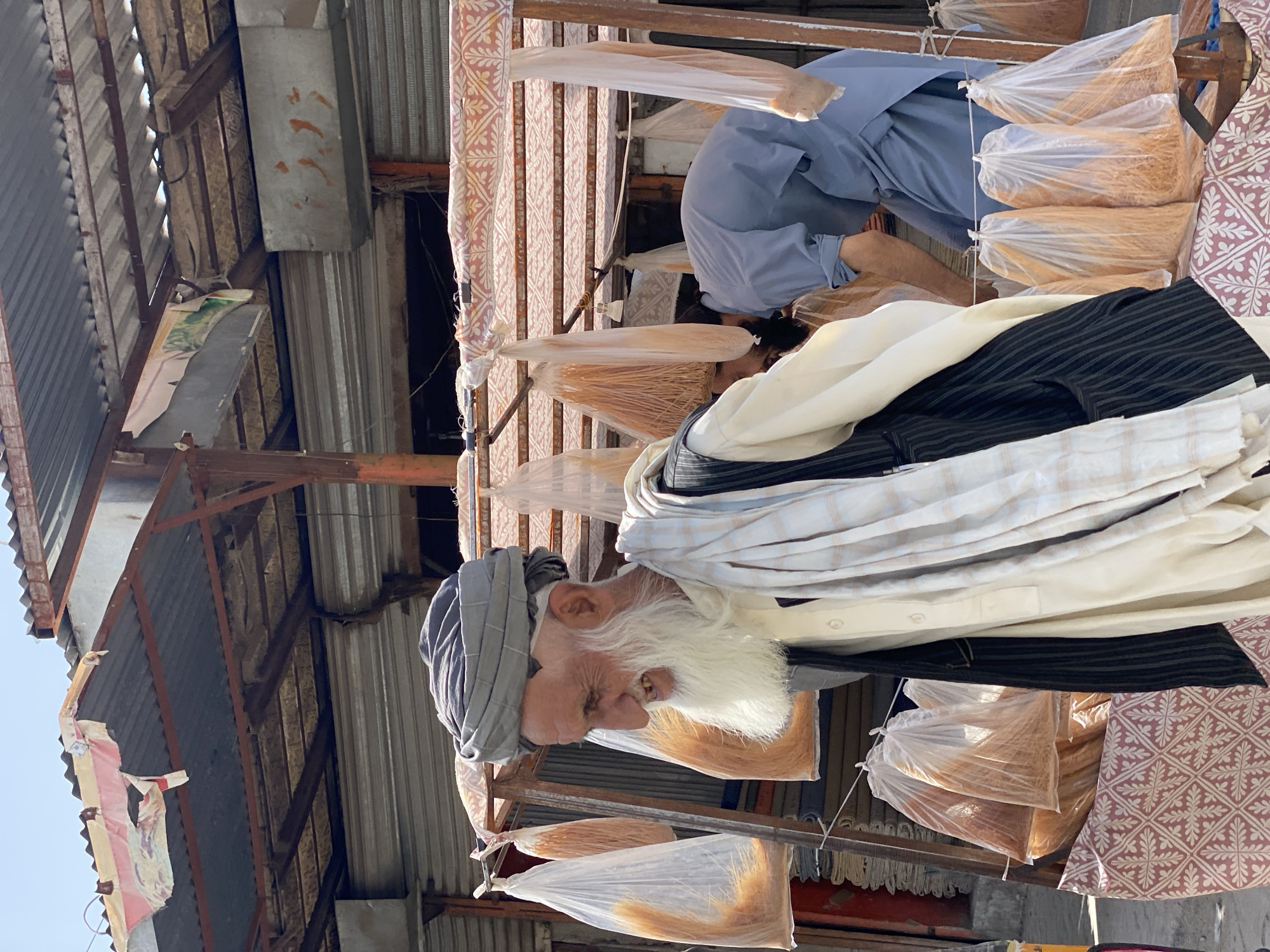 In Kandahar, Afghanistan, an elderly man with a long white beard and a grey turban stands in front of a market stall. He is wearing a dark vest over a light-colored shirt and a striped scarf. Behind him, another man in a blue tunic is bending over, possibly attending to the goods. The stall displays numerous transparent plastic bags filled with dried, light-brown strands, likely noodles or vermicelli. The market has a corrugated metal roof with some torn fabric coverings and a sign with Arabic script visible. The overall scene depicts a typical day at an outdoor market in Kandahar.