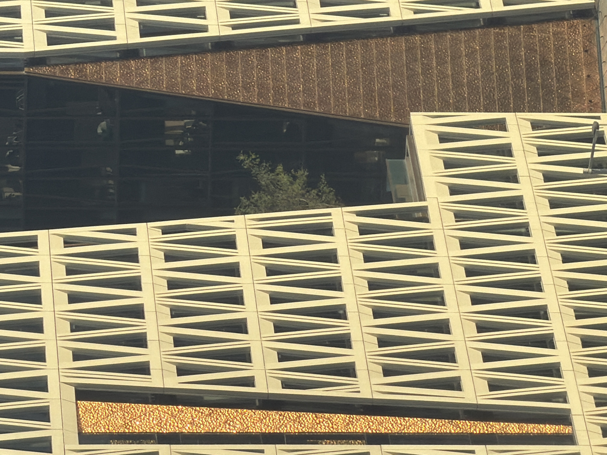 The image is a close-up, aerial view of modern skyscrapers. The building on the left has a beige facade with repeating triangular window frames. A vertical section of this building is clad in shimmering, textured golden material. The building on the right has a darker, possibly glass facade, revealing interior spaces with hints of office furniture and trees. A prominent, triangular section of this right building is covered in a dark, textured material that appears to be made of many small, reflective elements arranged horizontally. The two buildings are positioned to the left and right of a diagonal line, creating a sense of depth. The overall impression is one of architectural complexity and contemporary urban design.