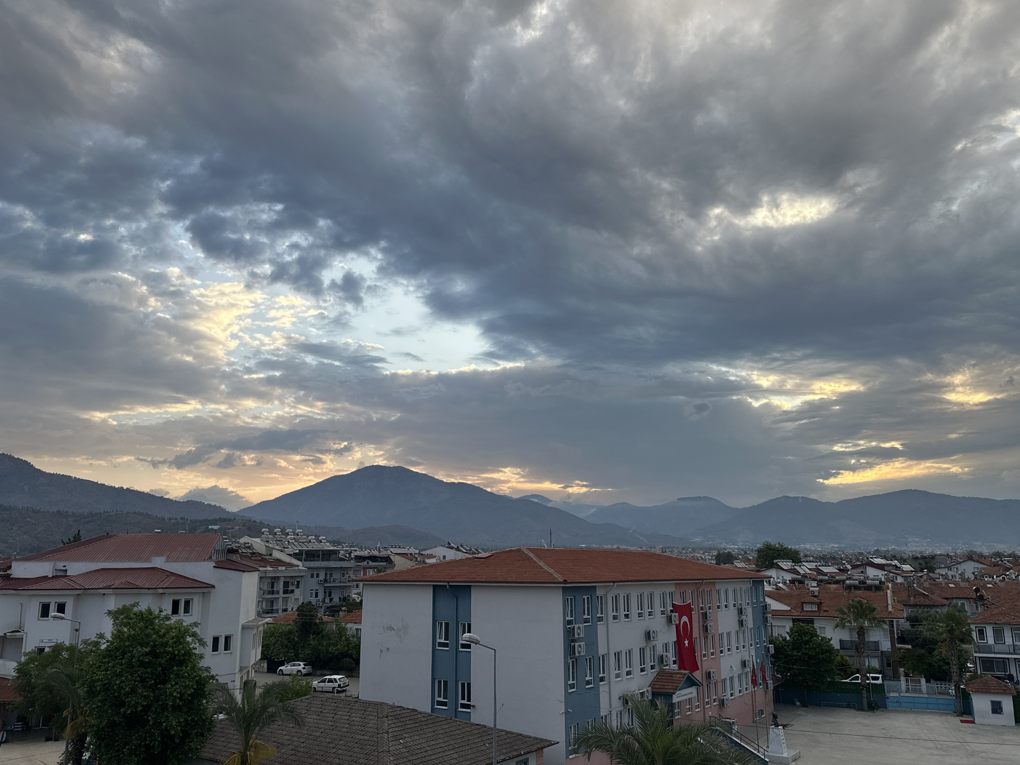 The image shows a landscape in Fethiye, Turkey, at dawn or dusk, under an overcast sky with hints of sunlight breaking through the clouds. The foreground is dominated by buildings with red tiled roofs and white or light-colored walls. One building features a Turkish flag hanging from its facade. Mountains are visible in the background, suggesting a hilly or mountainous terrain typical of the region. The area appears to be a residential or mixed-use neighborhood.