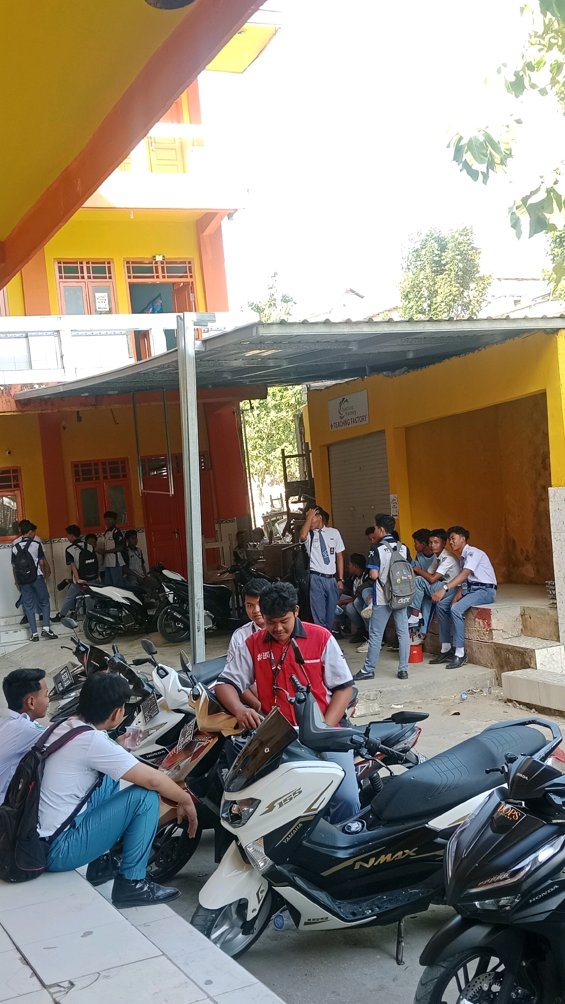 In Waru, Indonesia, a group of male students, dressed in school uniforms consisting of white shirts and blue trousers, are gathered outside a building. Several students are seen standing or sitting near motorcycles parked in front of a structure with an orange and yellow facade. Some of the students are holding bags or wearing backpacks. One student in the foreground wears a red vest over his uniform. The environment appears to be an outdoor area adjacent to a building, possibly a school or educational facility, with motorcycles prominently featured.