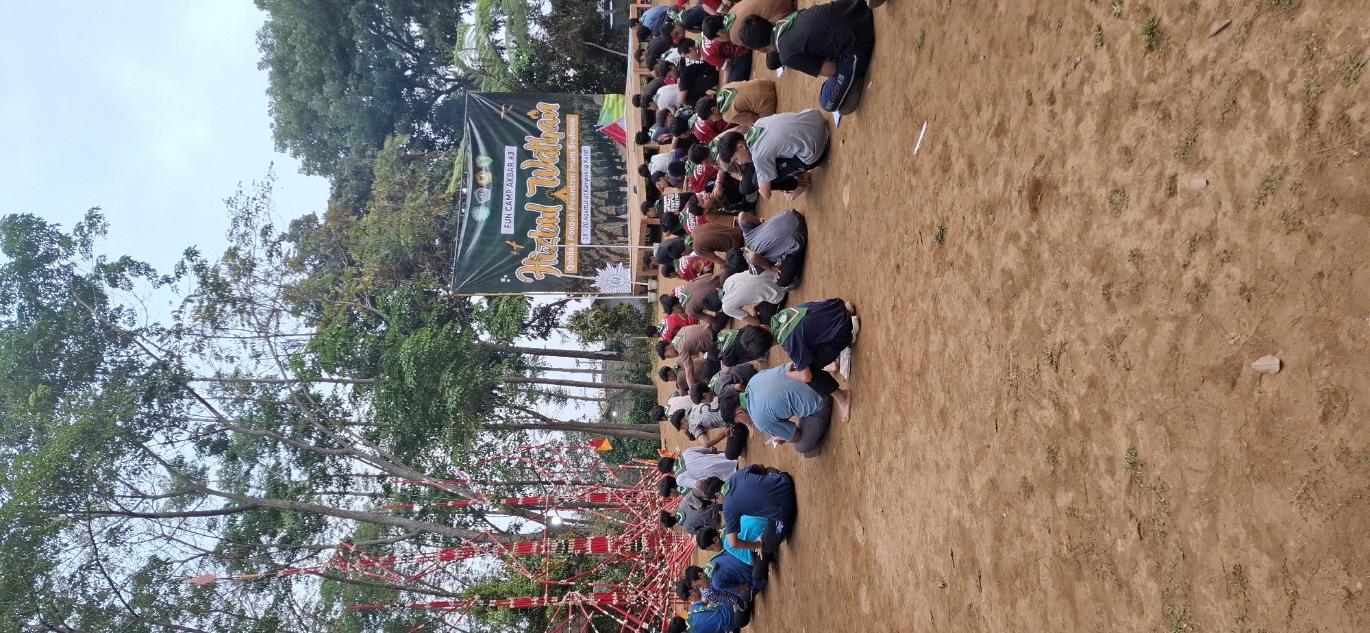In Puntukrejo, Indonesia, a group of approximately 50 young people, likely scouts, are seated on the ground in an outdoor setting. They are facing a banner that reads "FUN CAMP AKBAR #3 Hizbul Wathan Qobilan Pondok Pesantren Imam Syunodo 18-20 Agustus at Kampaeng Karet". The banner is green and features text and graphics. To the left of the banner is a tall, intricate structure made of red and white materials, possibly a decorative or symbolic installation. The ground is a reddish-brown dirt. Surrounding the area are trees, with foliage and branches visible against a pale sky. The attendees are mostly wearing uniforms or casual clothing, many with scout scarves. Some individuals appear to be looking towards the banner, while others are looking down or interacting with their peers. The overall atmosphere suggests an organized event or gathering, likely a camp or festival.
