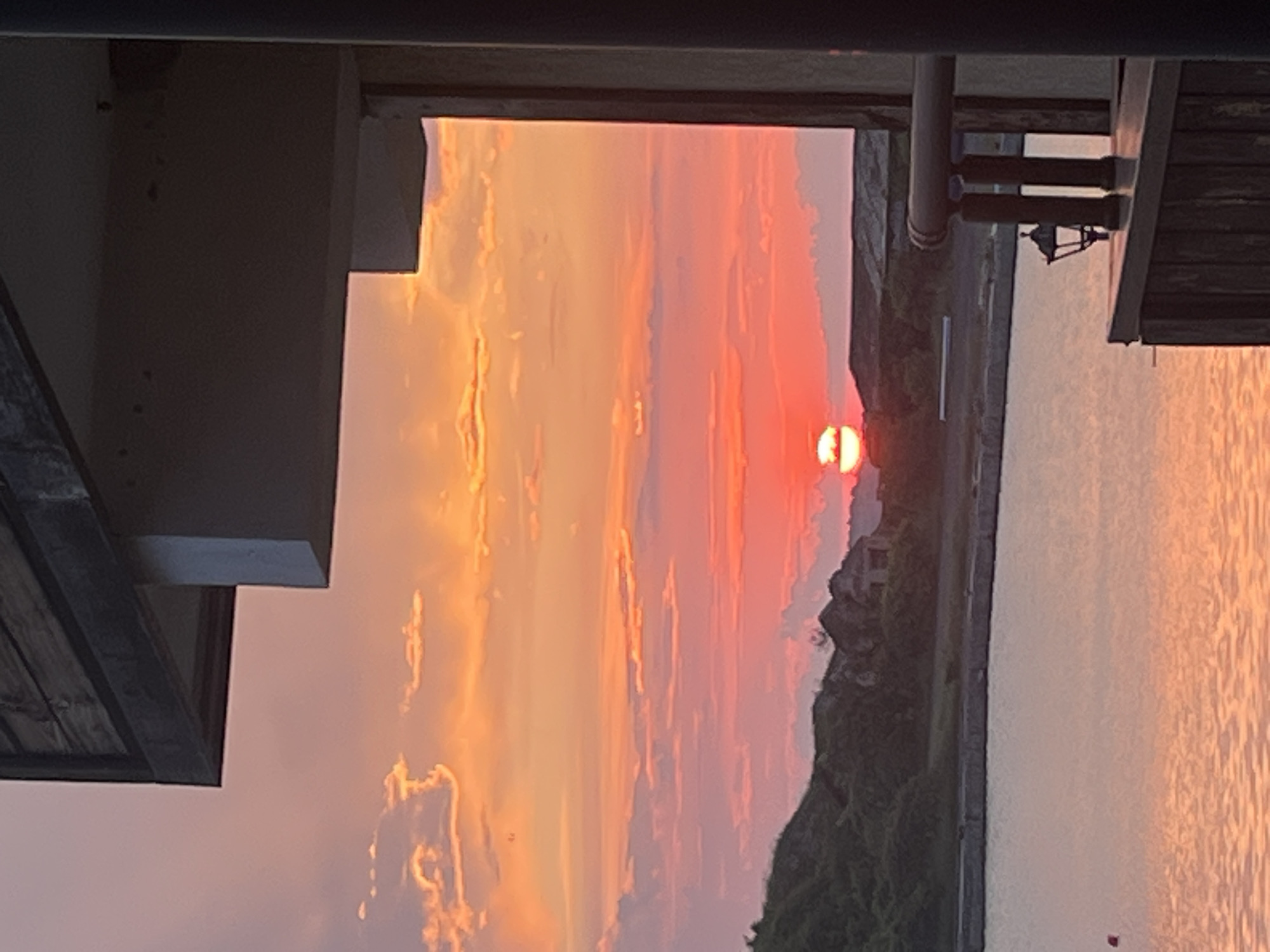 The image depicts a vibrant sunset over a body of water, viewed from what appears to be a structure or building.
The scene is dominated by the dramatic sky, showcasing a rich palette of orange, red, pink, and purple hues. Scattered clouds are illuminated by the setting sun, creating a dynamic display of color. The sun itself, a bright orange-red disc, is partially visible low on the horizon, casting a warm glow across the entire sky.
Below the horizon lies a calm body of water, likely an ocean or large lake, which beautifully reflects the vivid colors of the sky. In the mid-ground, across the water, a dark silhouette of a low mountain or landmass is visible, topped with some vegetation and possibly a distant structure.
The foreground is framed by dark, silhouetted architectural elements. On the left, a sturdy wall or pillar juts into the frame. Along the top, a horizontal beam or roof edge is present. To the right, a wooden structure, possibly part of a balcony railing or pier, is visible. Attached to this wooden structure are a dark, pipe-like object and a small, distinct silhouette of what appears to be a hanging lantern or light fixture.
There are no people visible in the image, suggesting a serene and undisturbed moment. The time of day is clearly sunset, as indicated by the low sun and intense sky colors. The weather appears clear and calm. There is no visible text. The overall impression is one of peaceful natural beauty and a captivating end to the day.