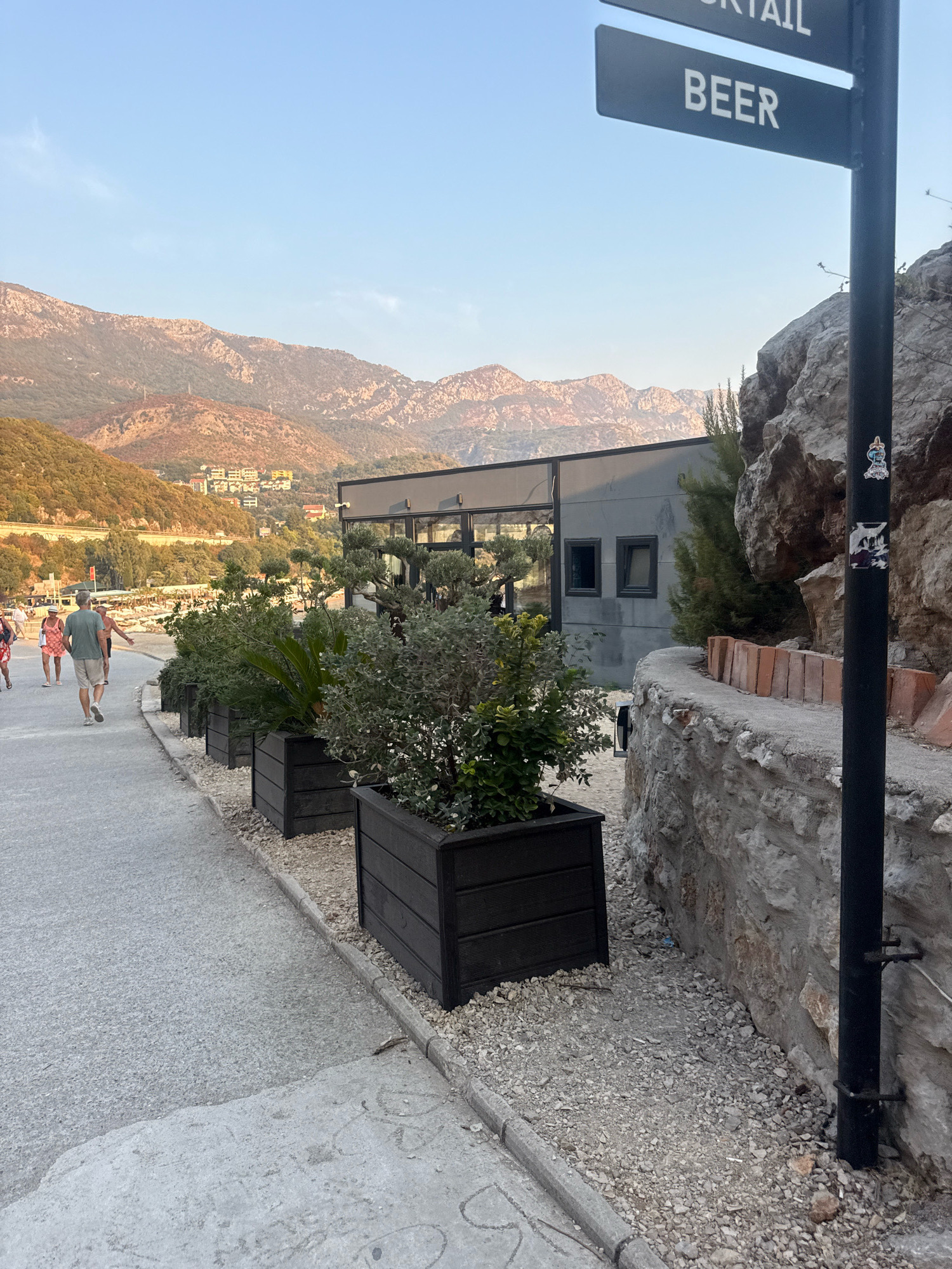 The image depicts a sunlit outdoor scene with a clear blue sky and hazy mountains in the background. In the foreground, a paved walkway runs alongside a row of dark planters filled with greenery. To the right, a rough-hewn stone wall curves along the path, adjacent to a black pole with two directional signs indicating "COCKTAIL" and "BEER." A modern building with large windows is visible behind the planters and the walkway. Several people are walking on the path in the mid-ground, dressed in casual summer attire. The setting appears to be a coastal area, possibly within Kamenovo, Montenegro, given the mountainous backdrop and the presence of a walkway suggestive of a promenade or beach access. The overall atmosphere is relaxed and summery.