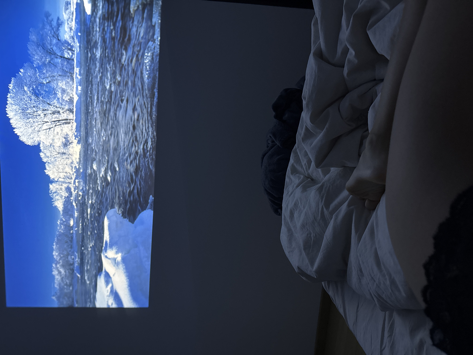 The image depicts a bedroom scene. A screen displaying a winter landscape with snow-covered trees and a flowing river is visible on the left side of the frame. On the right side, there is a bed with a white duvet, showing a portion of a leg, foot, and lace-trimmed garment. The overall lighting is dim, suggesting a nighttime setting. There is no indication in the image that can identify the location as the city of Warsaw, Poland.