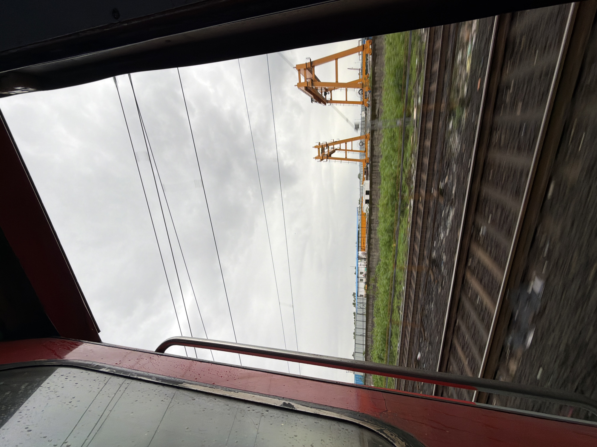 This image depicts a view from a moving train in Mira-Bhayander, India. The foreground is dominated by the interior of the train car, with a portion of a red and grey exterior visible on the left, featuring a window with water droplets. The train tracks are in sharp focus, showing multiple parallel lines receding into the distance. Beyond the tracks, a strip of green vegetation runs horizontally across the frame. In the mid-ground, two large, orange gantry cranes stand prominently against a cloudy, overcast sky. Behind the cranes, a row of structures, possibly warehouses or industrial buildings, are visible. The overall scene suggests an industrial or port area viewed during a train journey.