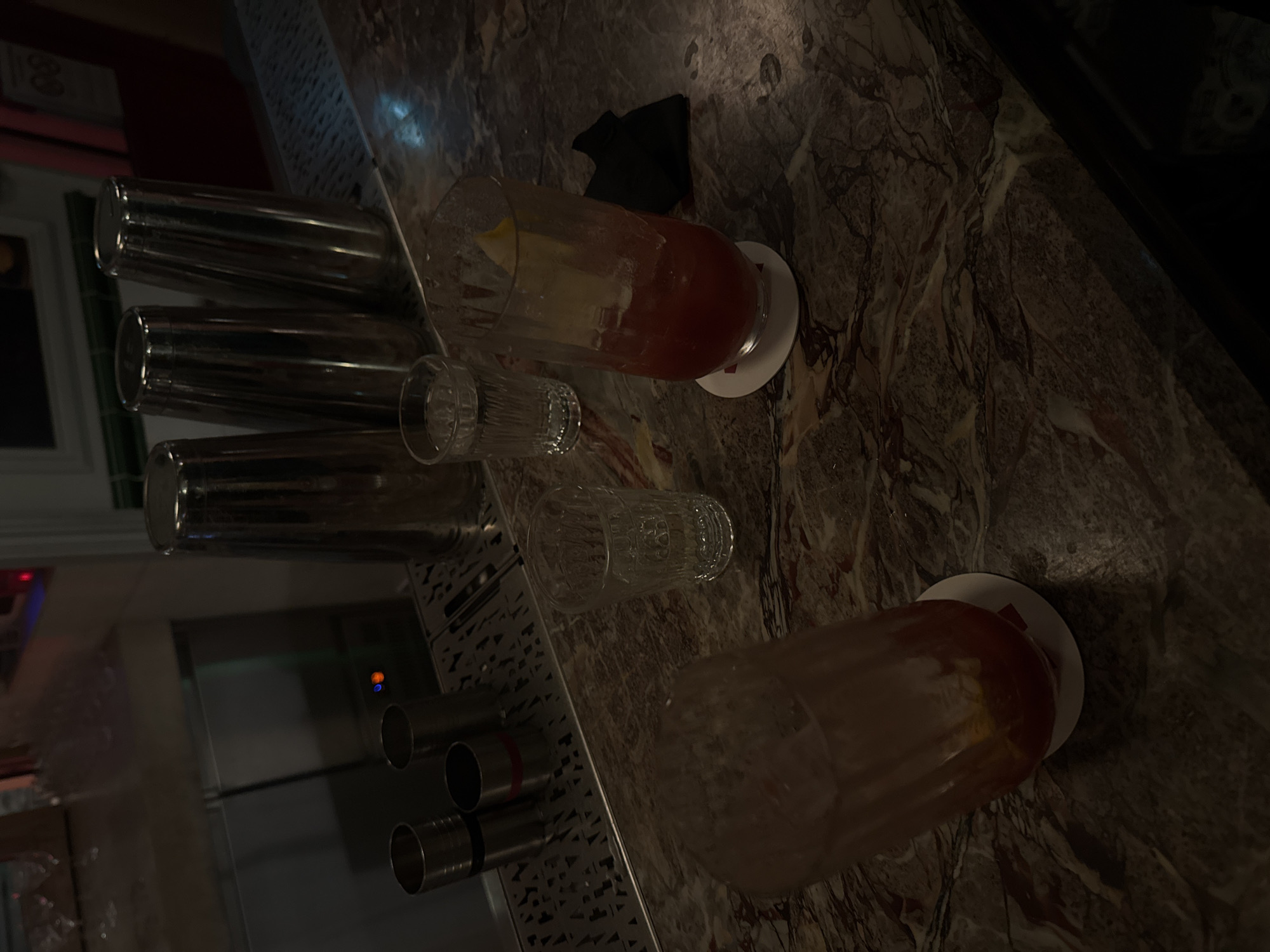 The scene depicts a dimly lit bar setting, likely in a sophisticated establishment. Two cocktails, one with a red hue and a lemon garnish, are resting on coasters on a marble countertop. A black napkin is near one of the cocktails. Several cocktail shakers, two small glasses, and bar equipment are visible on the bar's surface. The background shows a metallic structure, possibly part of the bar setup, and a glimpse of the interior. There is no indication of Paris, France.