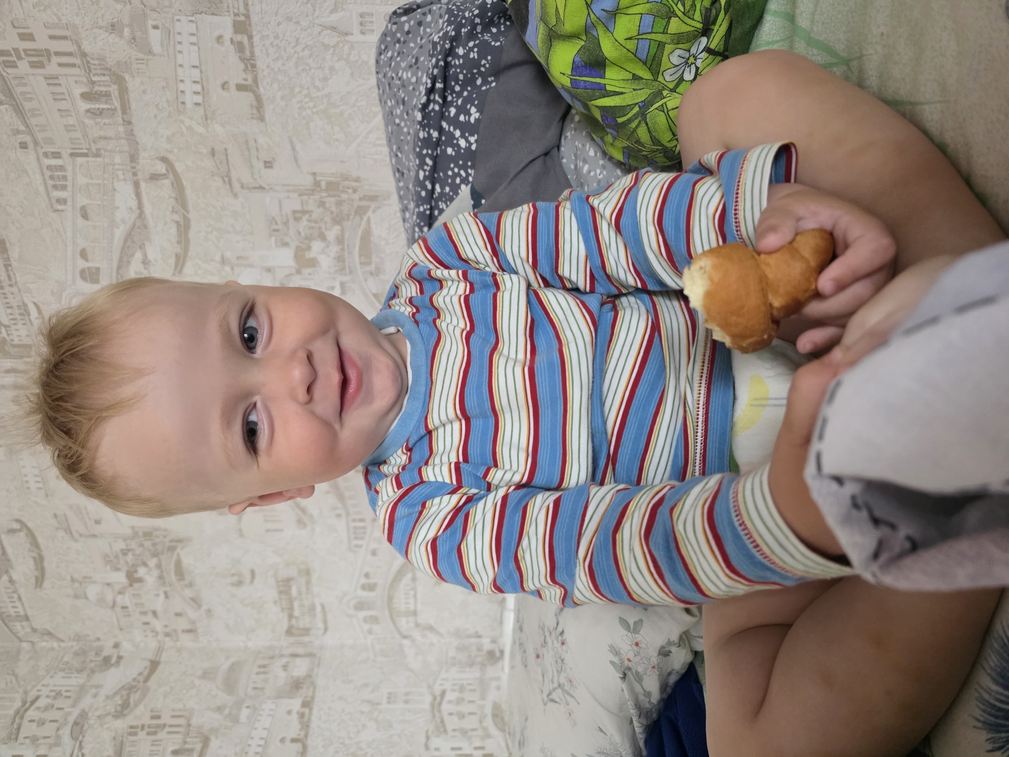 A young child, likely a toddler, is depicted indoors, looking directly at the camera with a wide, happy smile. The child has blonde hair and blue eyes and is wearing a long-sleeved striped shirt featuring red, blue, green, and white vertical lines. They are holding a small, light brown, crescent-shaped pastry, possibly a croissant or roll, in their right hand; part of it appears to have been eaten.
The child is seated or propped up on a bed or soft surface, surrounded by various patterned pillows and bedding. A green pillow with a leaf pattern is visible to the child's right, and a grey blanket with white specks is seen behind their left shoulder. Another white pillow with delicate floral patterns is partially visible near their legs. The background features light-colored wallpaper adorned with a subtle, muted pattern of buildings and architectural elements, suggesting an urban or classical motif. The lighting is bright, indicating an indoor setting during daytime. No text is visible in the image.