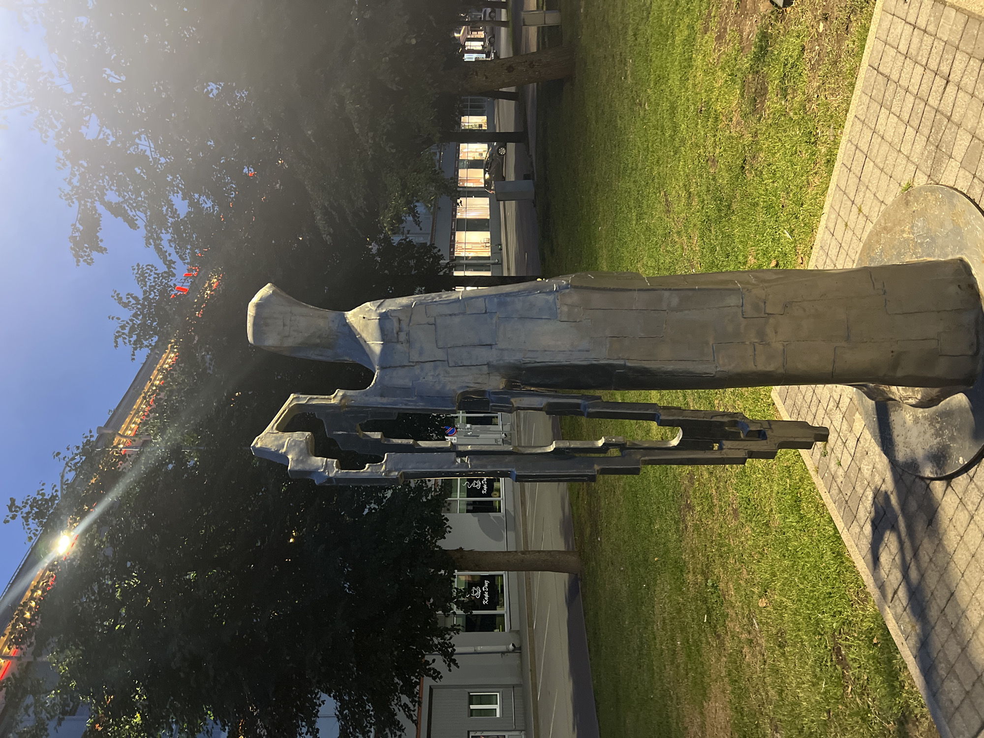 The image depicts an outdoor scene at dusk in Veliky Novgorod, Russia. In the foreground, a modern abstract metal sculpture stands on a paved area. The sculpture is metallic gray, with a textured surface composed of rectangular blocks. It has a tall, slender form with an intricate, geometric cutout section on its left side, and a more solid, cloaked figure-like shape on its right. A shadow of the sculpture is cast onto the patterned pavement.
Behind the sculpture, a well-maintained green lawn stretches towards a street lined with trees. In the middle ground, there is a building with large windows that are illuminated from within, suggesting it is a commercial establishment. The windows have white logos, one of which reads "Kafe Dug". Across the street, more trees are visible, and there are parked cars.
In the background, the upper portion of a building is visible, adorned with red decorative lights and illuminated by a bright streetlamp that casts a strong beam. The sky above is a deep blue, transitioning to the darkness of night. The overall lighting suggests artificial illumination from streetlights and the building, as well as the fading natural light of dusk. The scene appears tranquil and urban, with the sculpture serving as a focal point in a public space.