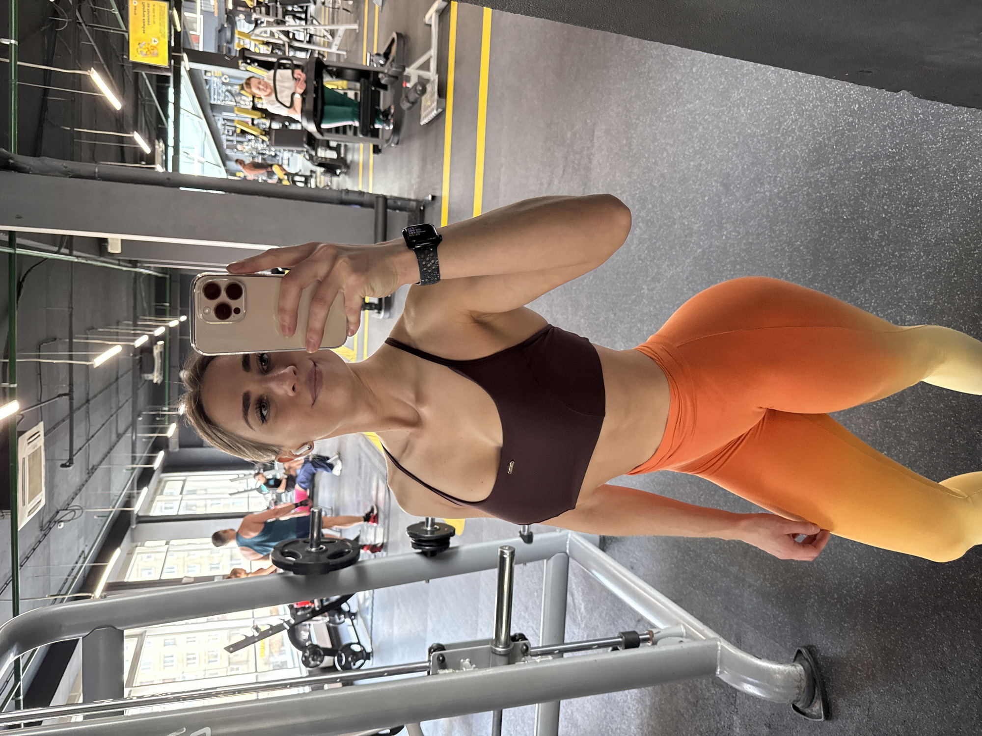 Here is a description of the image:
A woman is taking a selfie in a gym. She is wearing a dark sports bra and orange-to-yellow gradient leggings. She is holding a phone in her right hand and has a smart watch on her left wrist. In the background, there are gym machines and other people exercising. The location appears to be a fitness center in Saint Petersburg, Russia, as suggested by the view of buildings visible through the windows.