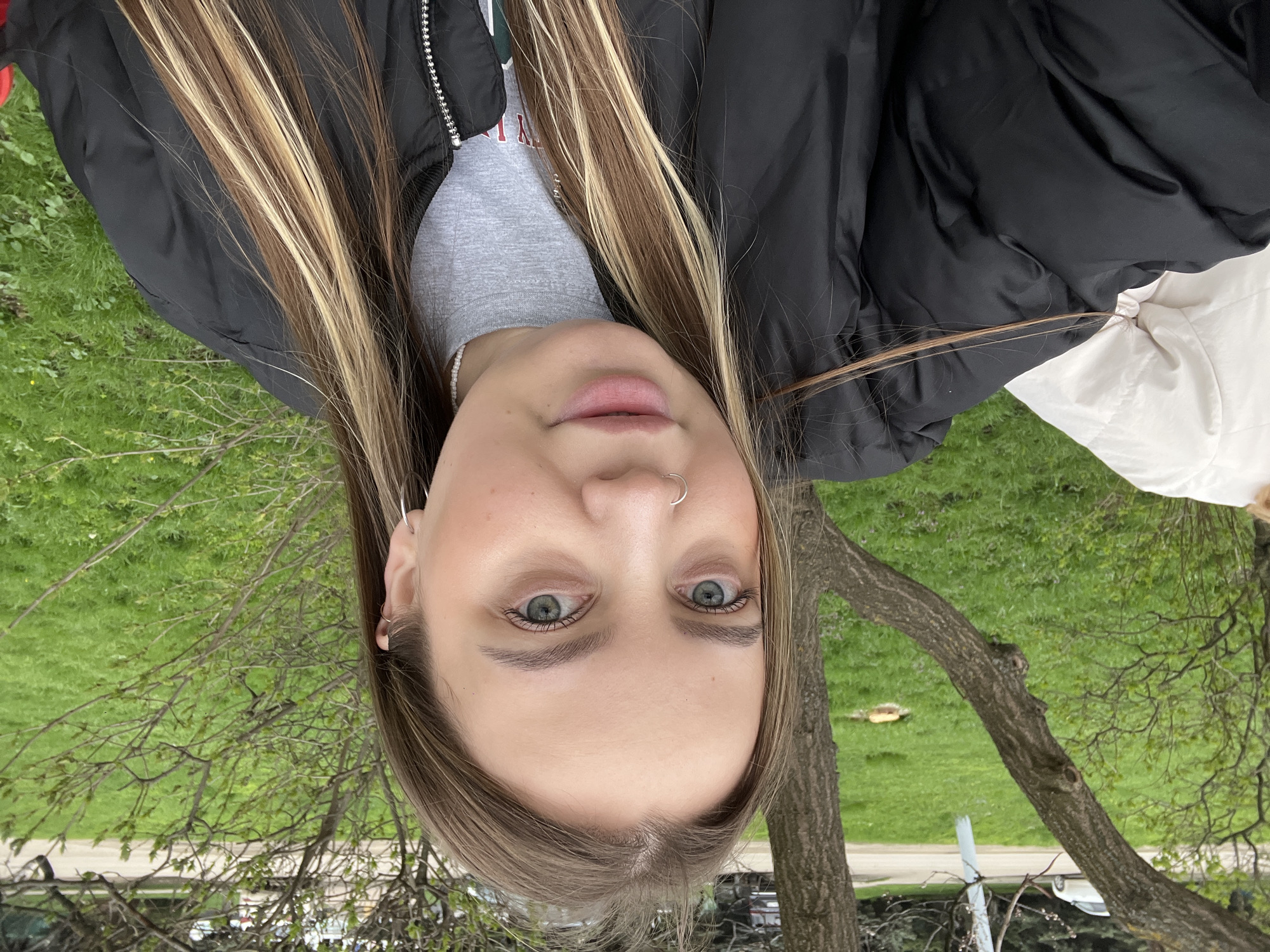 A woman with long, highlighted brown hair is facing the camera. She has green eyes and a nose ring. She is wearing a grey t-shirt and a black jacket. Behind her is a green lawn and a tree. The location is most likely outdoors in a park or green space. The image does not contain enough detail to determine whether the location is in Novopokrovskaya, Russia.