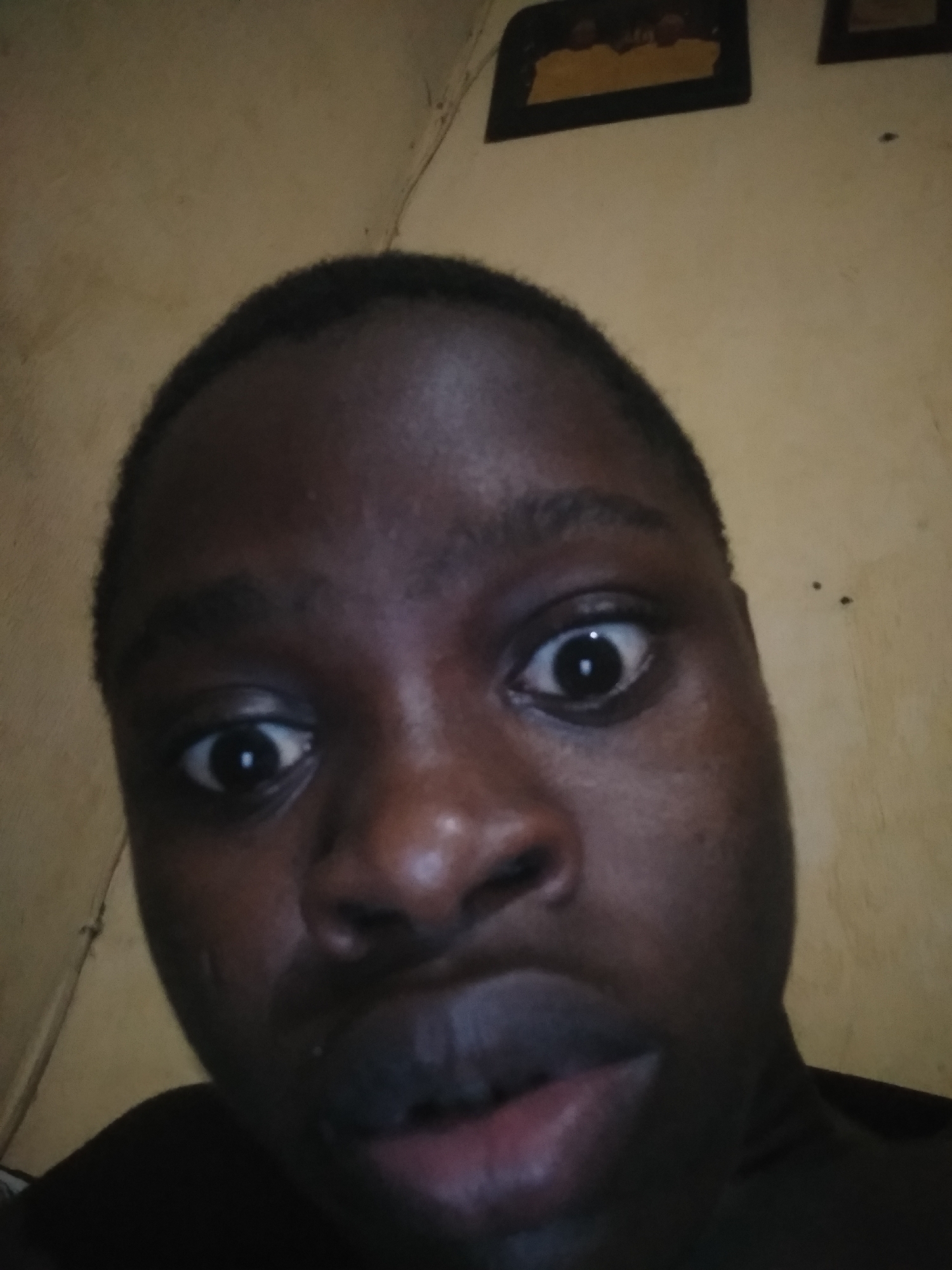 A close-up selfie of a young Black male with closely cropped hair. He has wide eyes and a slightly surprised expression. The background consists of a light yellow wall with a few dark framed pictures hanging near the top. The location is indicated as Unknown, Unknown.