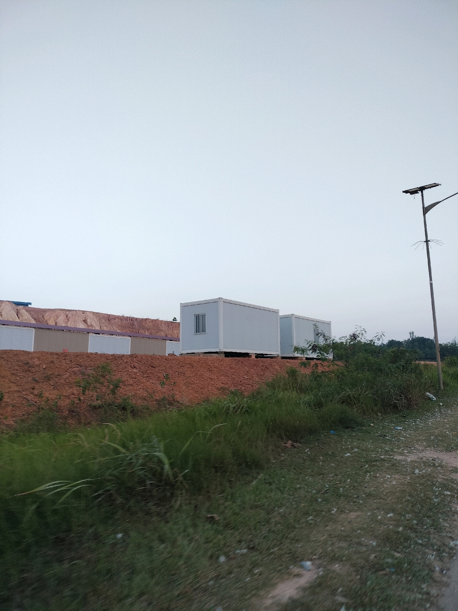 The image depicts a construction site in Batam, Indonesia, characterized by modular housing units and earthworks. In the middle ground, several white modular buildings are visible, appearing to be temporary accommodations or offices. These structures are placed on raised foundations. Behind them, a large embankment of reddish-brown soil suggests ongoing excavation or landscaping. The foreground is dominated by overgrown green grass and some scattered debris on a dirt path. A street light with a solar panel is positioned on the right side of the image, indicating a location that likely experiences regular power outages or aims for sustainable energy. The sky is overcast, casting a soft, diffused light over the scene, and the overall atmosphere suggests an early morning or late evening setting. There are no individuals visible in the frame.