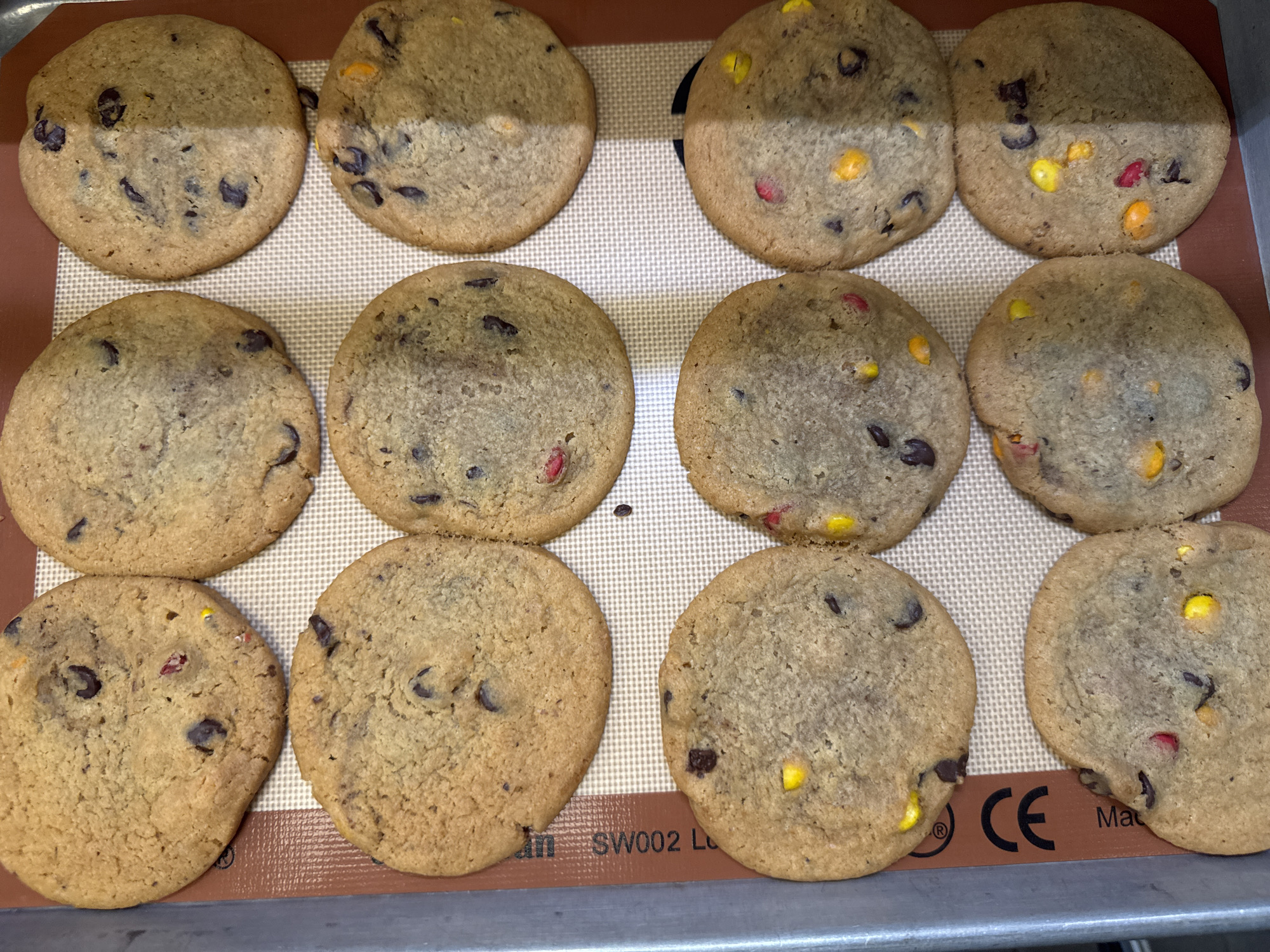 A dozen freshly baked cookies are arranged in rows on a tan baking mat. The cookies are round and flat with a slightly rough texture. They appear to be chocolate chip cookies with colorful candies mixed into the dough. Some cookies have dark chocolate chips, while others have visible red, yellow, and orange candies. The cookies are placed on a rectangular baking mat with a textured pattern. The mat is on a silver baking sheet. The cookies are arranged in three rows of four. The cookies in the first row are positioned towards the top of the frame, while the cookies in the third row are towards the bottom.