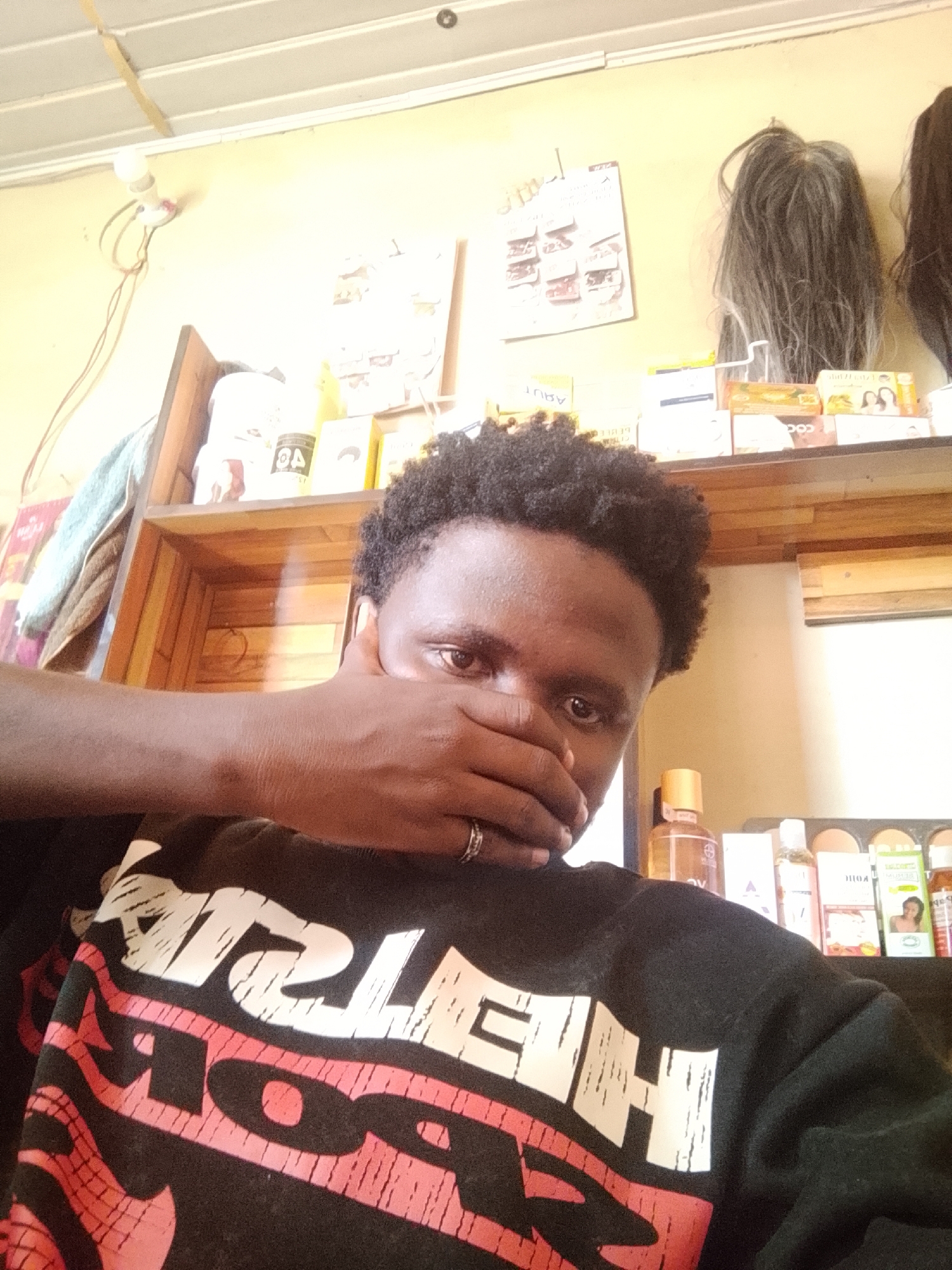 A young Black person is shown in a selfie, covering their mouth with their right hand. They have short, dark, curly hair and are looking directly at the camera with a serious expression. They are wearing a black sweatshirt with large white and red lettering across the chest. A silver ring is visible on the finger covering their mouth.
The background appears to be an indoor setting, possibly a shop or a room with shelves. The shelves are stocked with various items, including bottles, boxes, and what appear to be wigs displayed on the top shelf. Some of the boxes are labeled with text, but it is largely illegible. There is a light bulb with wires hanging from the ceiling in the upper left corner. The lighting suggests it could be daytime, but there are no clear indicators of the weather. The overall atmosphere is casual.