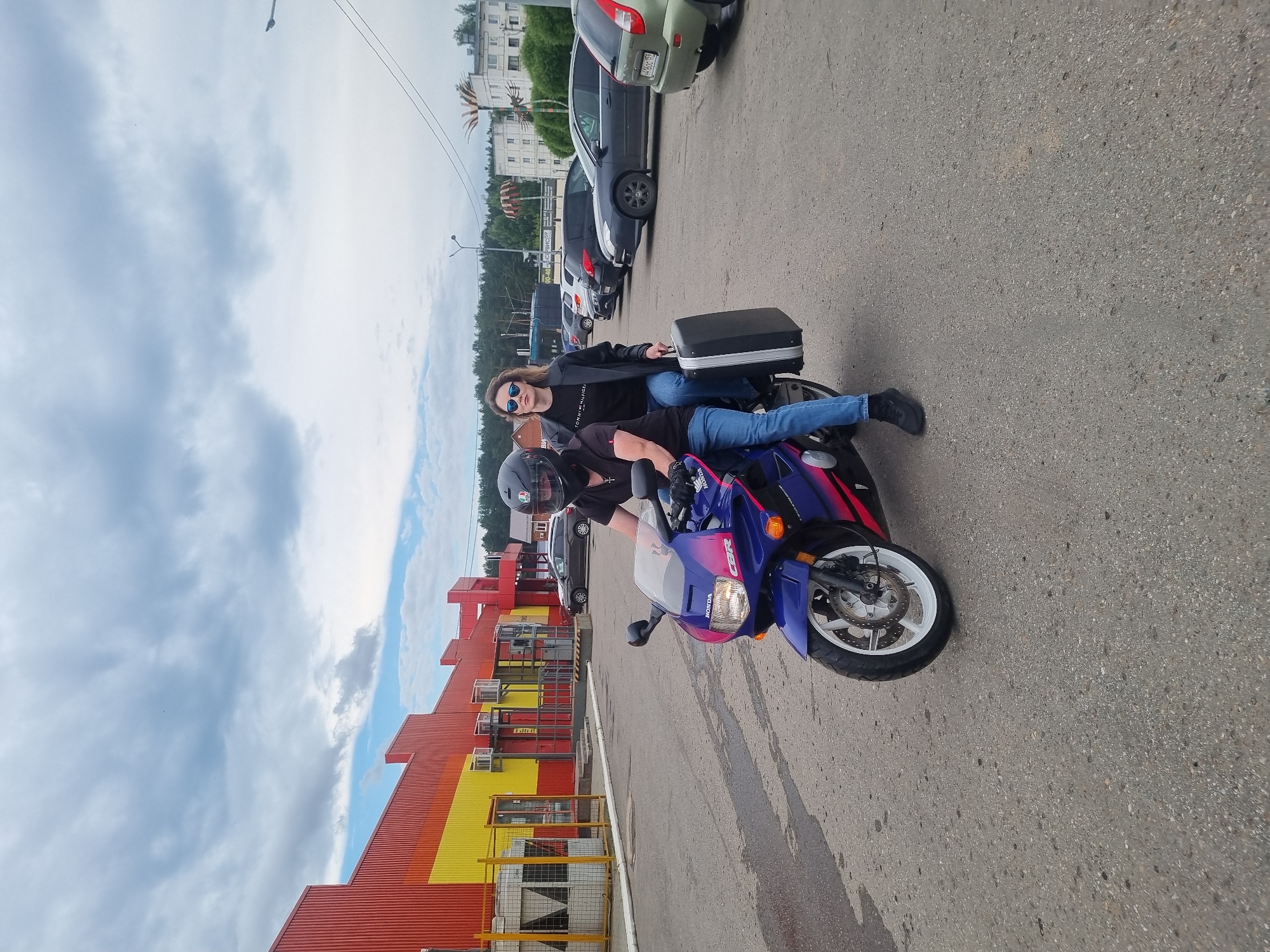 Two individuals are positioned on a blue and purple Honda CBR motorcycle in an outdoor paved area. The driver, wearing a black helmet, black t-shirt, and blue jeans, operates the motorcycle. The passenger, also in blue jeans, wears a black t-shirt with "TOMMY HILFIGER" visible and sunglasses. The passenger holds a black suitcase in their lap.
The background features a long, brightly colored building with red and yellow sections, appearing to be a commercial or industrial structure. Several parked cars are visible in the mid-ground. The environment includes a cloudy sky and a treeline in the distance, suggesting a location on the outskirts of a settlement. The scene is consistent with a location in the city of Chaikovsky, Russia, specifically an area with commercial buildings and parking lots adjacent to residential or natural surroundings.