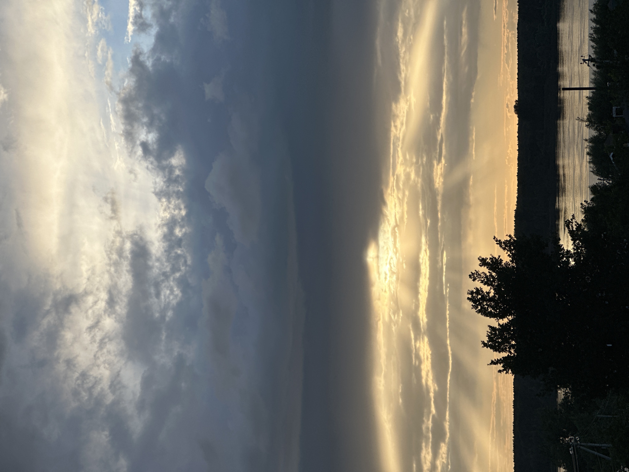 This is a vertical, low-angle outdoor shot of a dramatic sky over a body of water, likely a river or lake, at dusk. The sky is divided by a line of dark, heavy clouds running diagonally from the top left to the bottom right. To the left of this line, the sky is a mix of lighter, wispy clouds illuminated by the sun, with patches of blue sky visible. To the right of the dark clouds, the sky is a golden-orange hue, with the sun's rays breaking through in distinct beams, creating a spotlight effect.
The foreground features a dark silhouette of trees and foliage along the right edge of the frame. Beyond the water, a dark, tree-covered shoreline is visible, creating a stark contrast with the bright sky. The water itself reflects the colors of the sky, with shimmering ripples indicating a slight breeze. A utility pole with wires is faintly visible on the right side of the image, near the water's edge.
The overall mood is serene and awe-inspiring, with the dramatic lighting of the sunset creating a sense of grandeur. The time of day is clearly evening, with the fading sunlight and the colors in the sky indicating sunset. The location is Chaikovsky, Russia, suggested by the presence of a river or lake and the type of vegetation. There is no visible text in the image.