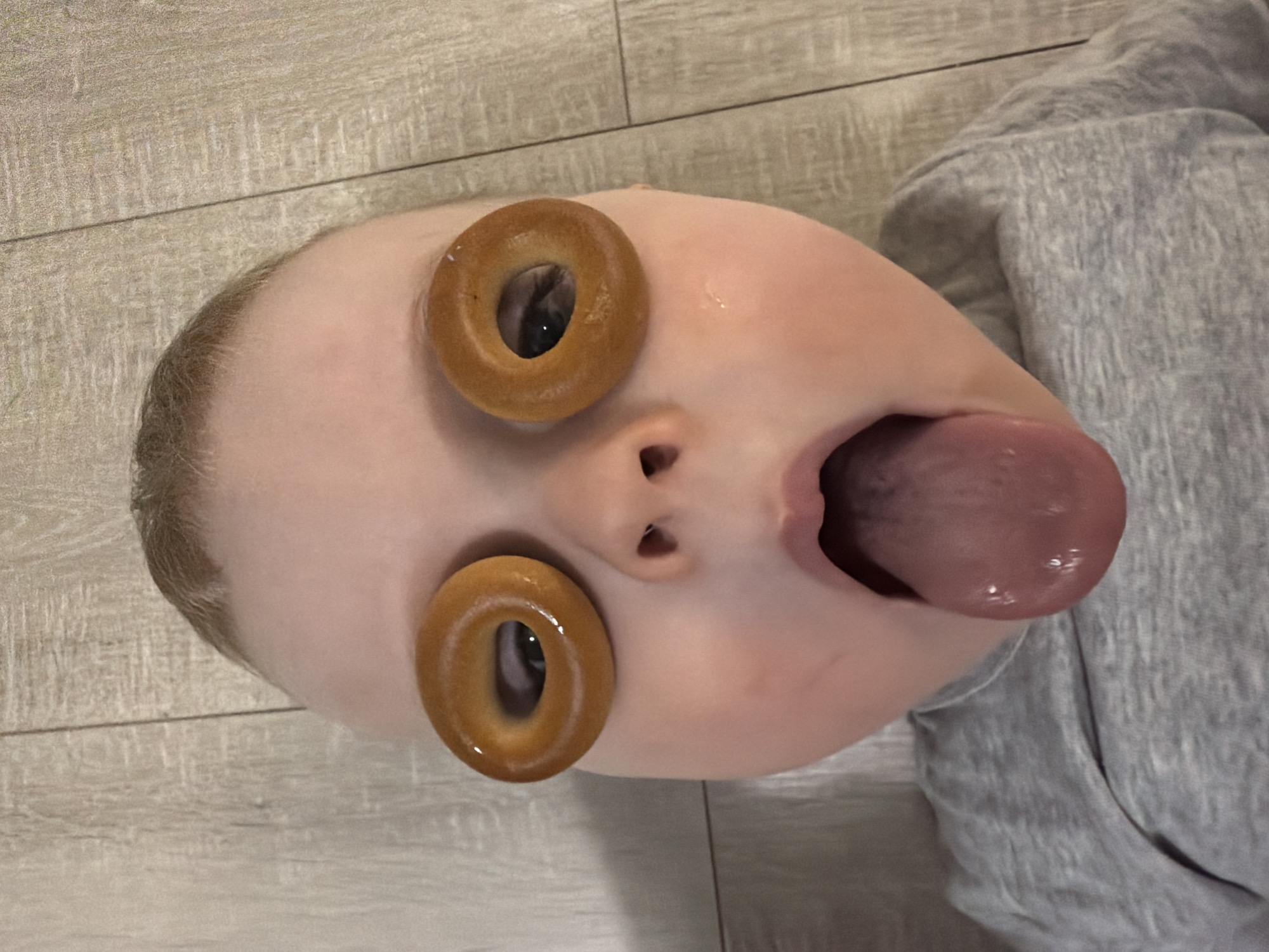 A baby lies on their back on a light wood-patterned surface. The baby's face is prominently featured, with two bagels placed over the eyes. The baby's mouth is open, and the tongue is extended. A gray sweatshirt covers the baby's upper body. The city of Votkinsk, Russia, is not discernible in this image.