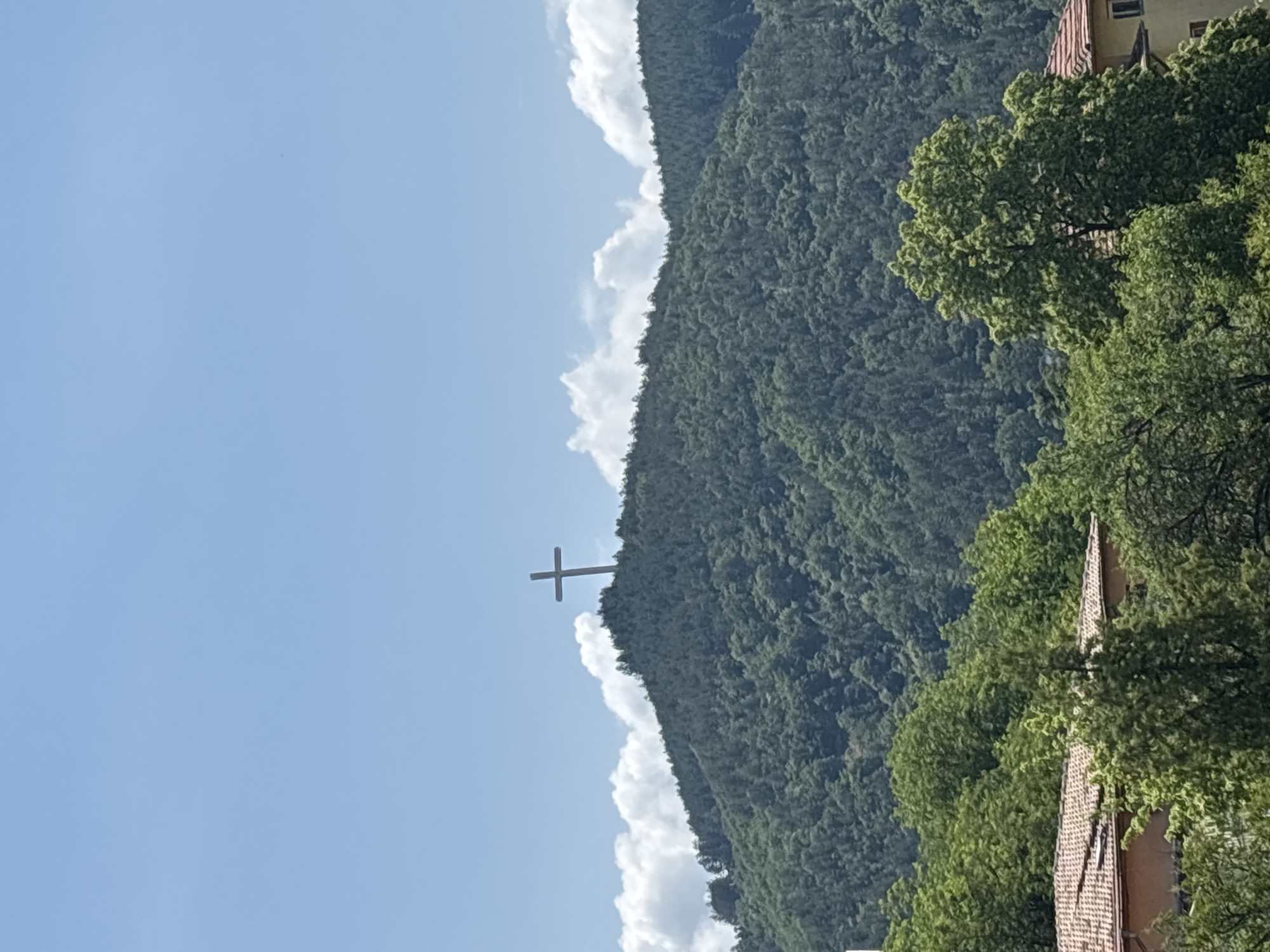 This image captures a panoramic view of a densely forested mountain or hill under a bright blue sky, with a prominent cross monument on its peak. The scene is oriented as if looking up towards the mountain, suggesting the photo was taken from a lower elevation, likely within a town.
The main subject is a large, verdant mountain covered in lush green trees, stretching across the middle and right side of the frame. Towards the left side of the mountain's crest, a tall, stark white cross stands out against the sky, positioned just above the tree line. Along the mountain's ridge, several white, fluffy cumulus clouds are visible, creating a soft contrast with the deep green of the forest and the blue sky. The upper left portion of the image is dominated by the clear, azure sky, indicating good weather.
In the lower right corner of the foreground, parts of buildings with distinctive reddish-brown tiled roofs and green treetops are visible, suggesting the presence of a settlement or residential area from which the photograph was taken.
The time of day appears to be daytime, likely late morning or early afternoon, given the bright, even lighting and clear sky. The weather is sunny and fair with scattered clouds. There is no visible text in the image. Based on the provided context, this is a view of the iconic Blagoevgrad Cross, located on Varosha hill, overlooking the city of Blagoevgrad, Bulgaria.