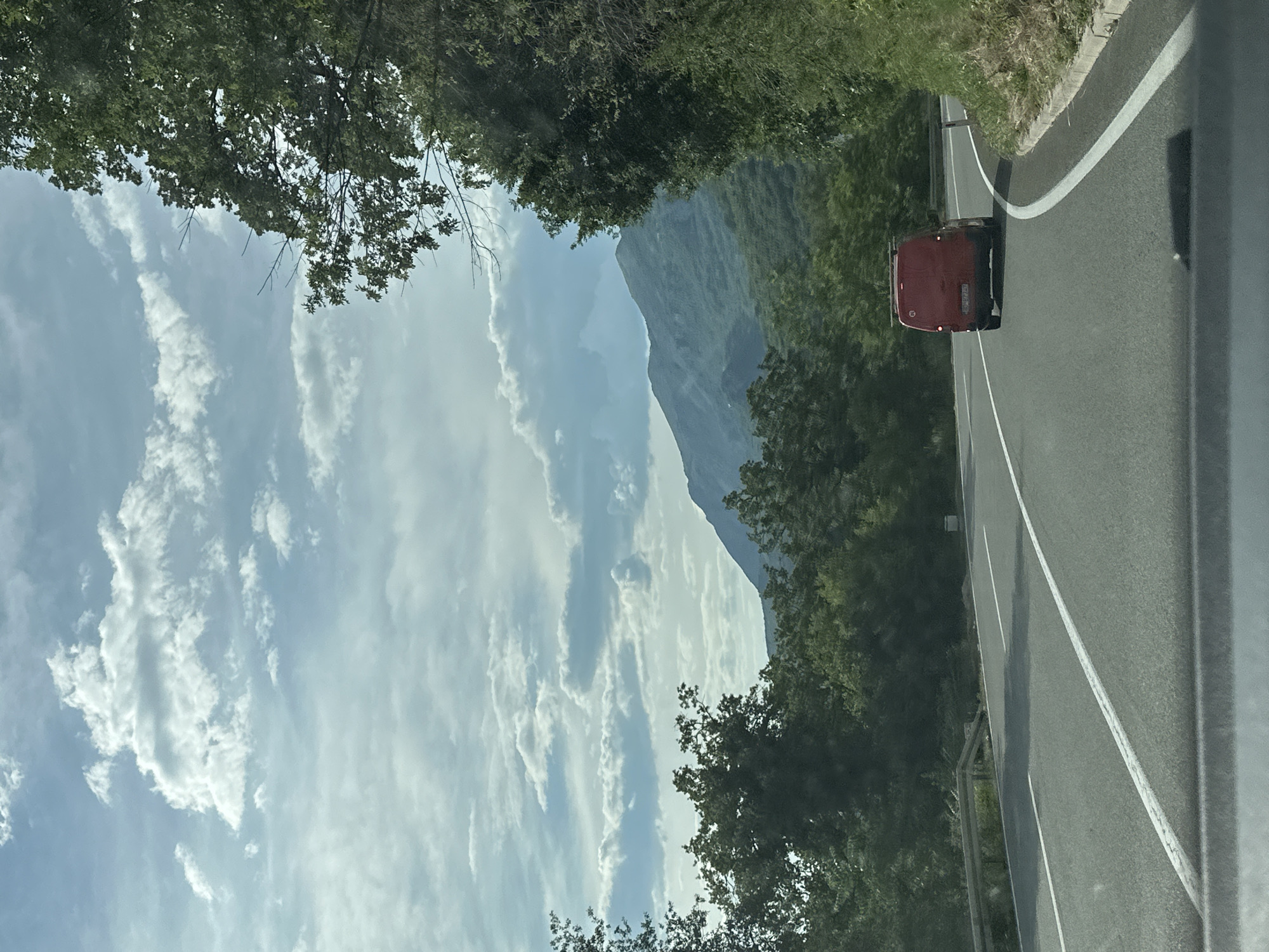 Captured from a moving vehicle on a road near Golubovići, Montenegro, the image depicts a landscape dominated by a mountain range under a partly cloudy sky. The road, marked with white lines, curves to the right. A red van travels on the road. Trees line both sides of the road, partially framing the mountain view.