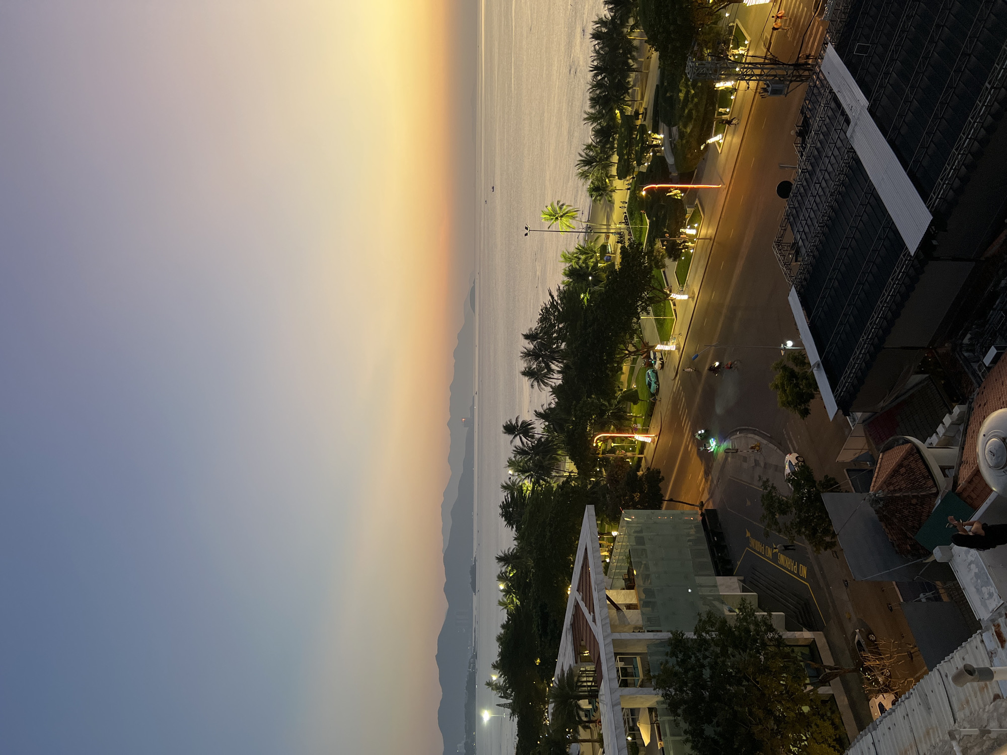 The image captures a serene coastal scene in Nha Trang, Vietnam, during the golden hour as the sun begins to set. The horizon is painted with a gradient of soft blues and warm oranges, with the vast expanse of the sea meeting a distant, hazy mountain range.
In the foreground, a modern building with a white facade and tinted glass is visible, possibly a hotel or resort, with balconies and outdoor seating areas. Adjacent to it is a street lined with lush palm trees and other tropical foliage, illuminated by streetlights and the warm glow of the setting sun. A few cars and a motorcycle are seen on the road, indicating some level of activity in the city.
Further back, a park area with manicured trees and pathways is visible along the beachfront, dotted with people enjoying the evening. The beach itself is largely empty, with gentle waves lapping at the shore. The overall atmosphere is one of tranquility and natural beauty, characteristic of coastal Vietnamese cities.