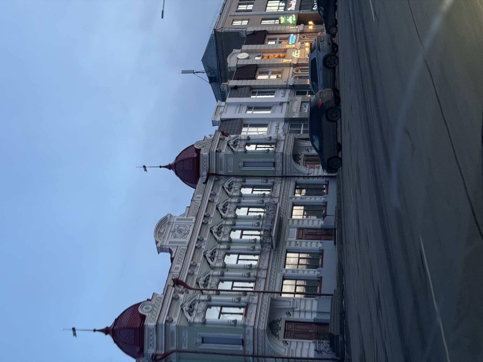 The image shows a street scene in Sarapul, Russia, featuring an ornate building with red domed roofs, elaborate decorative trim, and multiple windows. Several cars are parked along the street in front of the building. The sky is a pale blue-gray, suggesting it is dusk or dawn. Other buildings, some with lit signage, are visible further down the street. The perspective indicates the photo was taken from the street level.