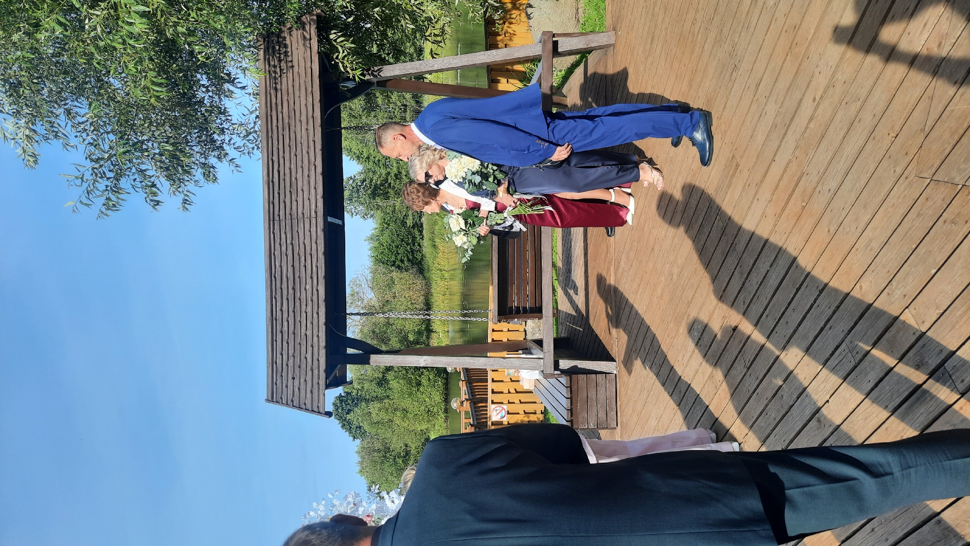 The image depicts an outdoor scene under a clear blue sky. In the foreground, a person in a dark suit is partially visible from the back, looking towards a group of people standing on a wooden deck. This group includes three women and one man. Two women are holding bouquets of white flowers. The man is wearing a blue suit. Behind them, a wooden swing set is attached to a thatched roof structure. The deck overlooks a calm body of water, likely a lake or river, with trees and foliage lining the opposite bank. The overall setting appears to be a park or a lakeside venue, possibly for an event. The location is identified as Жодзішкаўскі сельскі Савет, Belarus.