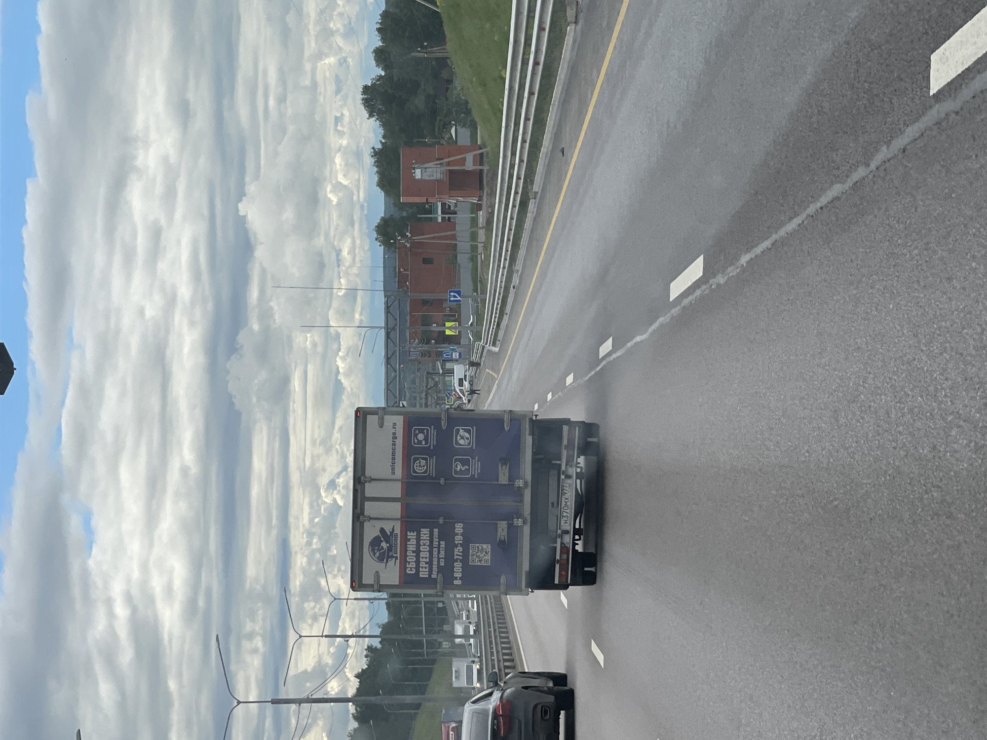 The image depicts a highway scene in what appears to be Taraskovo, Russia. The road is wet, possibly due to recent rain. A large truck, identified by its Russian license plate, is traveling ahead. The truck's back door features commercial graphics and contact information, including a phone number beginning with "8-800." The sky is overcast with layered clouds. Along the roadside, a metal guardrail is visible, with greenery and what appears to be a red brick structure in the background. Other vehicles are also present on the road.