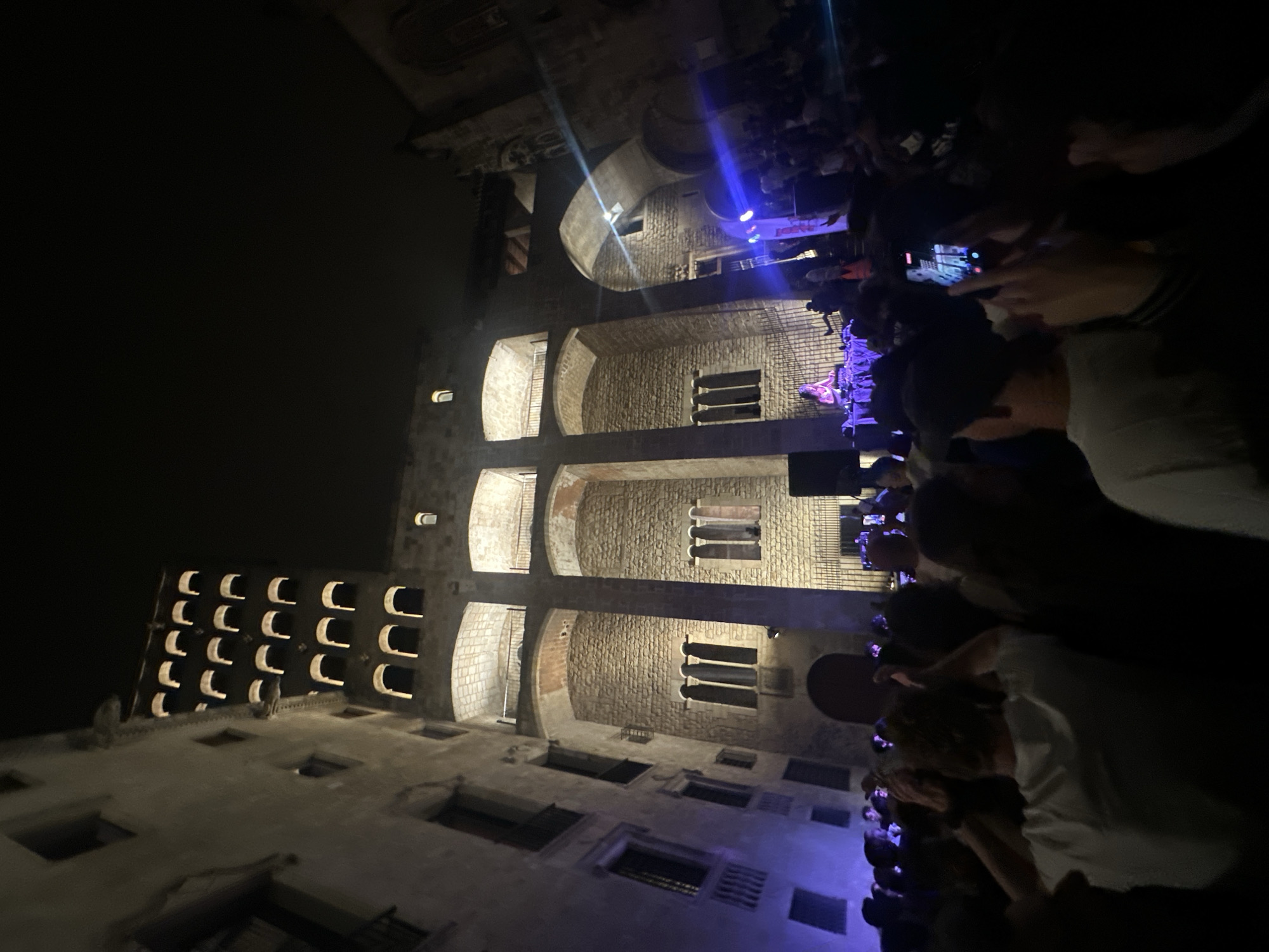 A night-time scene depicts a crowd facing a medieval stone structure, likely a castle or significant building. The crowd is dense, with many individuals holding up phones, presumably recording or photographing an event or performance occurring on the building's elevated levels. The stone structure features arched openings and recessed windows. The lighting is a mix of ambient light from the structure itself, some blue concert lighting, and the glow from screens on mobile devices. The city of Barcelona, Spain, is the location of this scene.