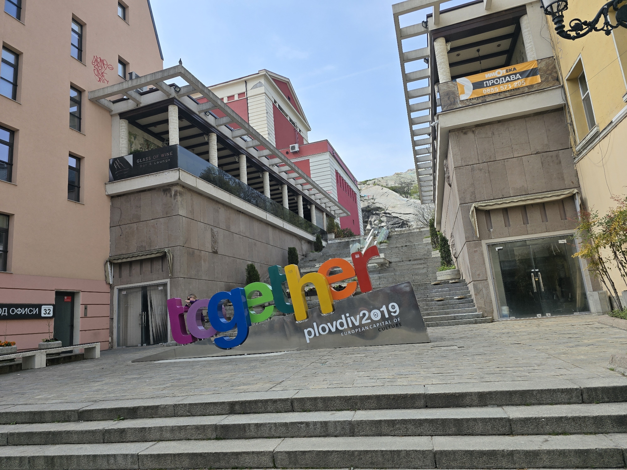 The image shows a cityscape, likely in Europe, featuring an art installation in the foreground. The installation spells out "together" in large, colorful letters, placed on a grey base with the inscription "plovdiv2019, EUROPEAN CAPITAL OF CULTURE." The installation is located on a stone-paved square or plaza, in front of a series of stone steps leading up a hill.
The buildings in the background vary in color and architectural style. To the left is a pink building with multiple windows and a restaurant or bar called "GLASS OF WINE" on the upper floor. To the right is a yellowish building, featuring a banner advertising real estate for sale, "ИМОТЕКА ПРОДАВА," with a phone number.
The sky is blue with a few clouds, suggesting a clear day. Overall, the image depicts a blend of historical architecture and modern art in a European city setting.