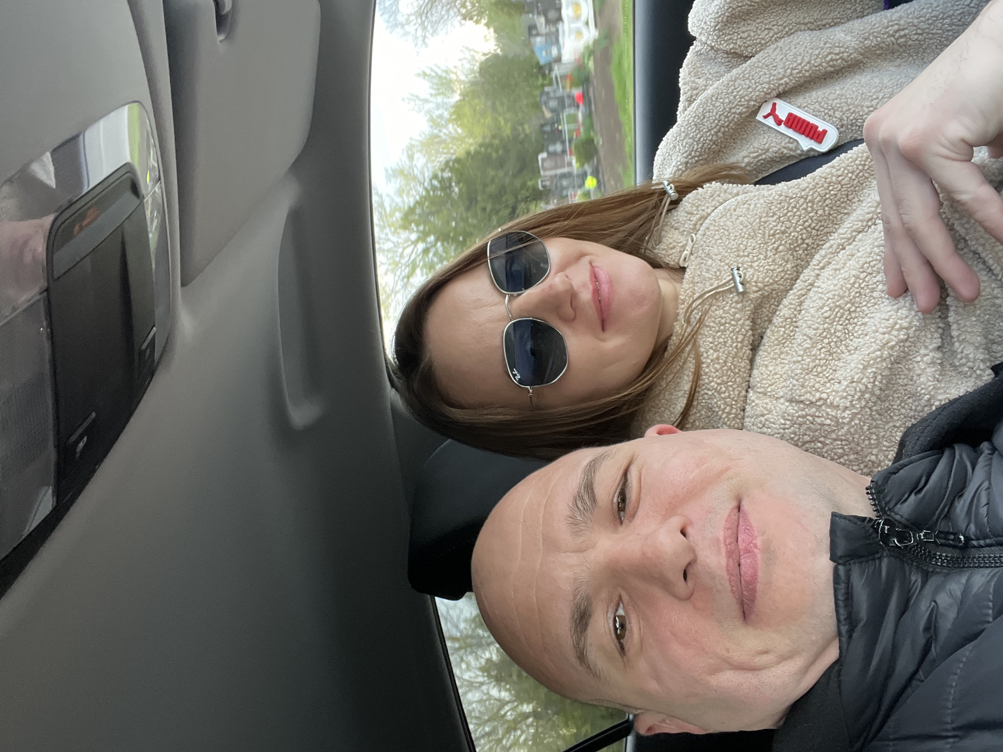 The image is a selfie taken inside a car, featuring two individuals, a man and a woman, with an outdoor scene visible through the window. The photograph is rotated 90 degrees counter-clockwise.
The woman is in the upper portion of the frame, smiling subtly with light pink lipstick. She has long brown hair and wears dark sunglasses, with "Ray-Ban" visible on the left lens. Her attire includes a cream-colored, textured, fuzzy jacket (sherpa-like material). A distinctive red and white accessory, shaped like a speech bubble with the "Puma" logo and brand name, is pinned to her jacket. Her left hand is visible, positioned near her chest.
Below her is the man, who is bald and looks towards the camera with a slight smile. He is wearing a black puffer jacket, with a partial view of its metallic zipper.
The interior of the car is primarily grey, with parts of the ceiling and an overhead console visible, showing a light or button panel. Through the window on the woman's side, an outdoor setting is visible, characterized by green trees and numerous white or light-colored structures that strongly resemble gravestones or markers, indicating a cemetery or burial ground in the background. The lighting suggests it is daytime. Both subjects appear relaxed.
Visible text includes "Ray-Ban" on the woman's sunglasses and "Puma" on the accessory attached to her jacket.