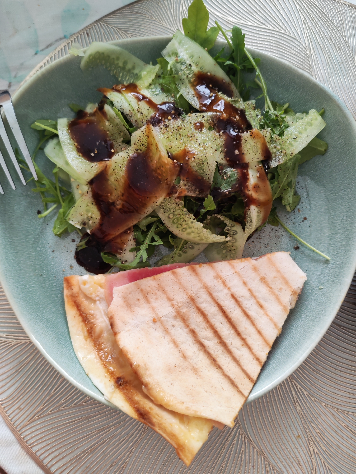 A close-up overhead shot shows a light blue ceramic bowl filled with a salad and a piece of grilled flatbread with cheese and a slice of ham. The salad consists of arugula and thinly sliced cucumber, dressed with a dark, glossy balsamic glaze and sprinkled with black pepper. The grilled flatbread is cut into a triangle and rests at the bottom right of the bowl, revealing melted cheese and a pink slice of ham. A fork is partially visible on the left side of the bowl. The bowl is placed on a textured placemat with a geometric pattern. The overall lighting suggests it is daytime. This image is not geographically specific to Izhevsk, Russia; it is a general food photograph.