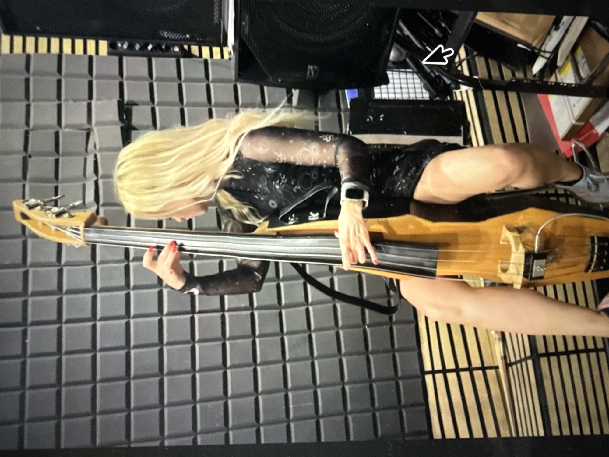 A woman with long blonde hair is playing a cello or upright bass. She is wearing a black outfit with sheer, patterned sleeves and a short black skirt. Her left hand is on the fingerboard of the instrument, and her right hand appears to be near the bridge. She has bright red nail polish. A black watch is visible on her left wrist.
The background consists of a wall covered in grey acoustic foam panels, arranged in a grid pattern. To the right of the woman, there are vertical wooden slats, possibly part of a stage or studio setup. Above, large black speaker cabinets are visible. The lighting suggests an indoor setting, likely a music studio or performance space. The overall mood seems focused and engaged, consistent with someone performing or practicing. No text is visible in the image.