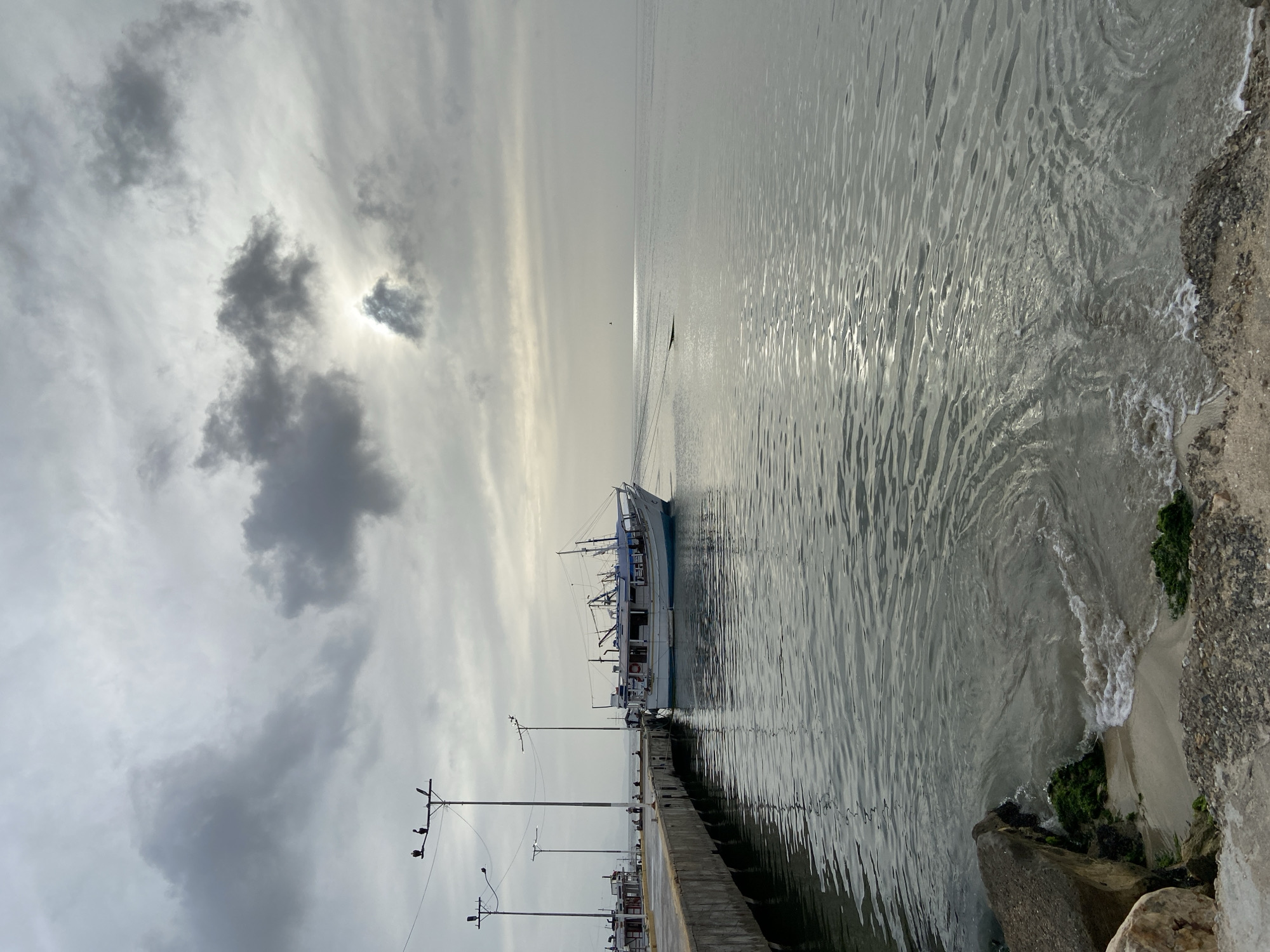 This image depicts the waterfront in the city of Juan Griego, Venezuela, under a cloudy sky. A white and blue fishing boat is docked at a concrete pier, with its reflection visible in the calm water. The pier is equipped with tall light poles, two of which have birds perched on them. Further down the pier, other smaller boats and several people can be seen. The water's surface is rippled, reflecting the subdued light filtering through the clouds. The foreground shows a small section of sandy shore with rocks and seaweed. The horizon is marked by the distant sea meeting the overcast sky, with the sun faintly visible behind a cloud formation.